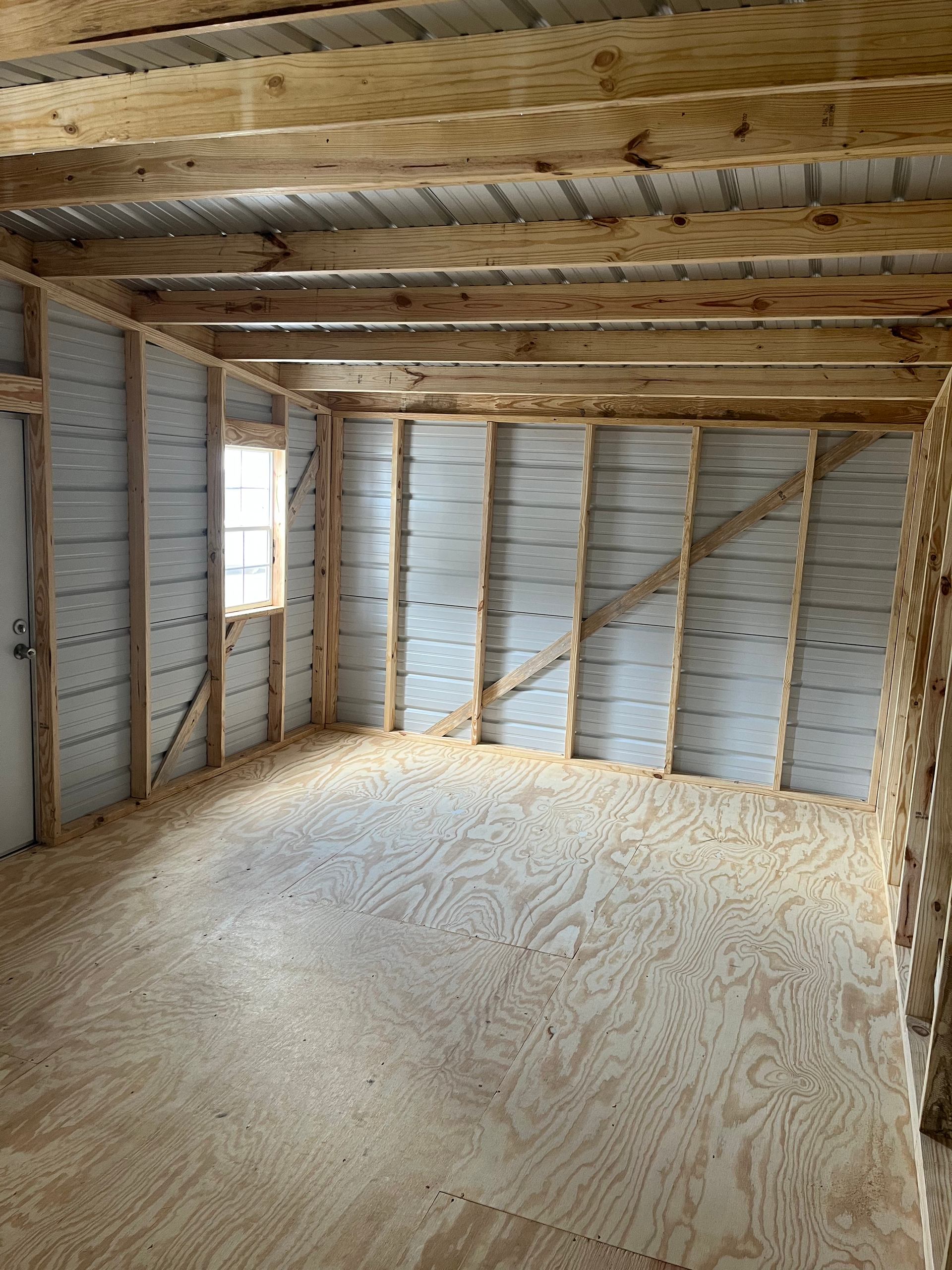 Empty unfinished room with exposed insulation, plywood floor, and wood ceiling beams.