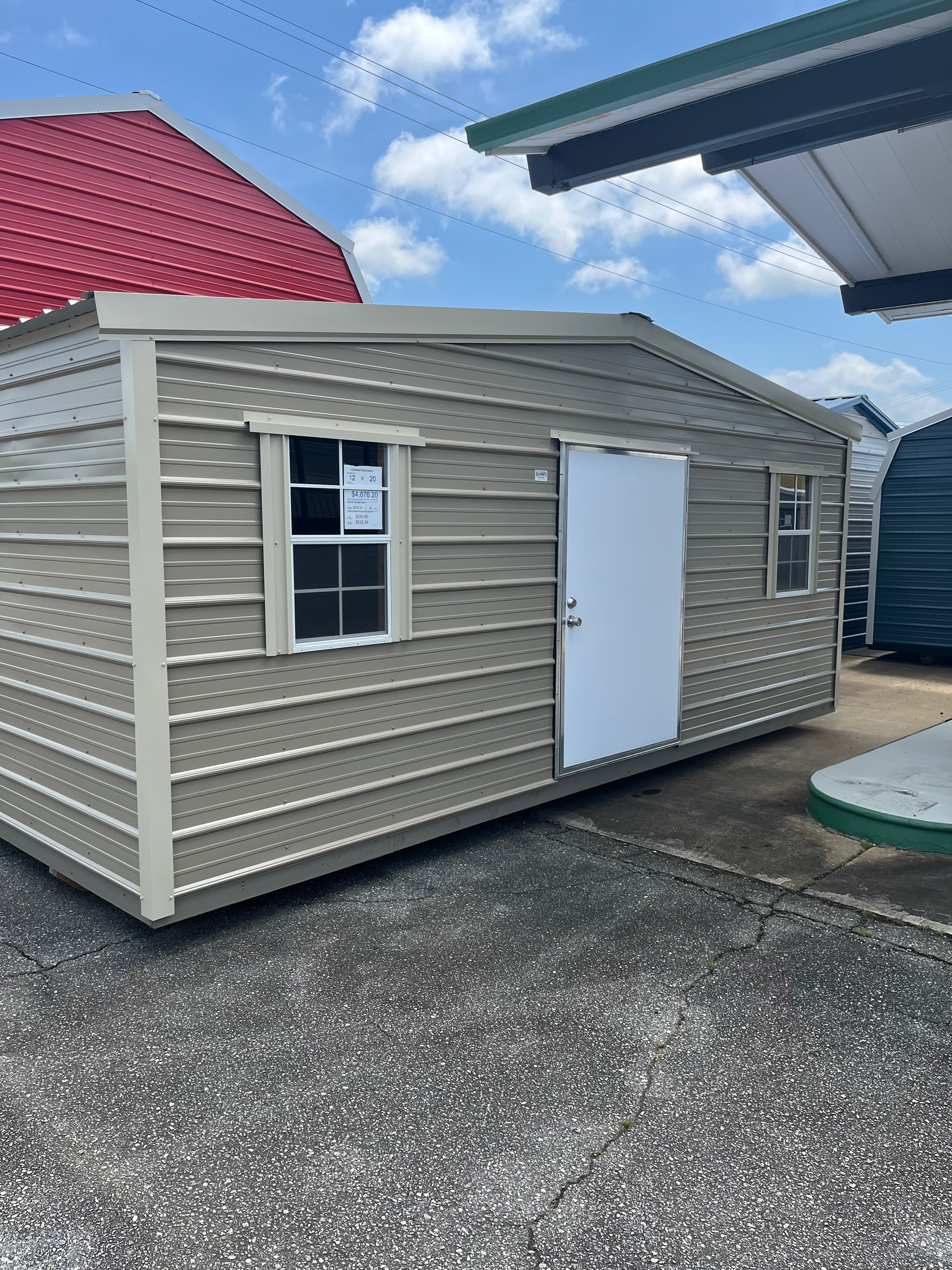Small beige shed with a white door and window on a gravel lot under a blue sky.
