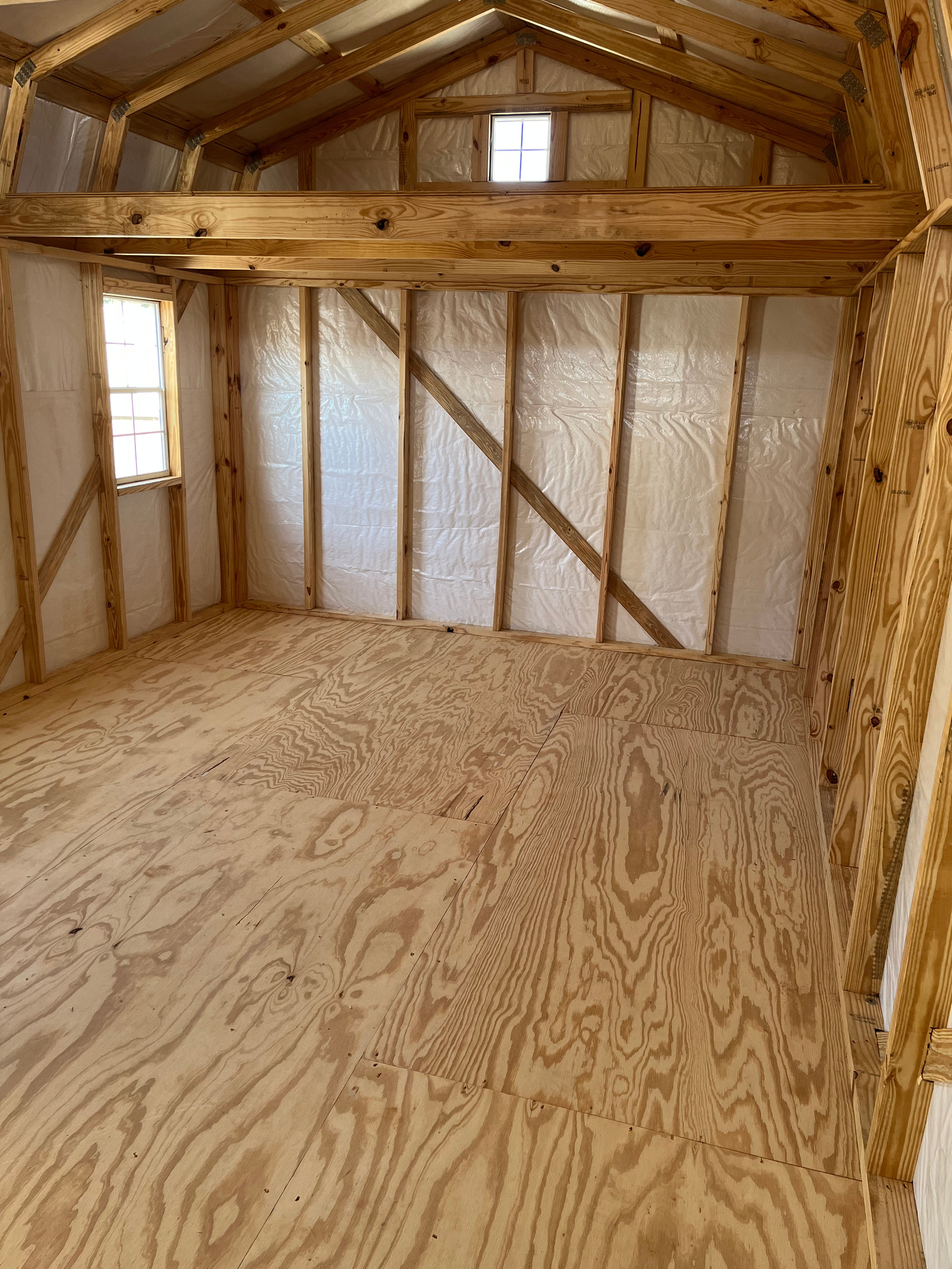 Empty unfinished wooden room with exposed studs, plywood floor, and small high window.