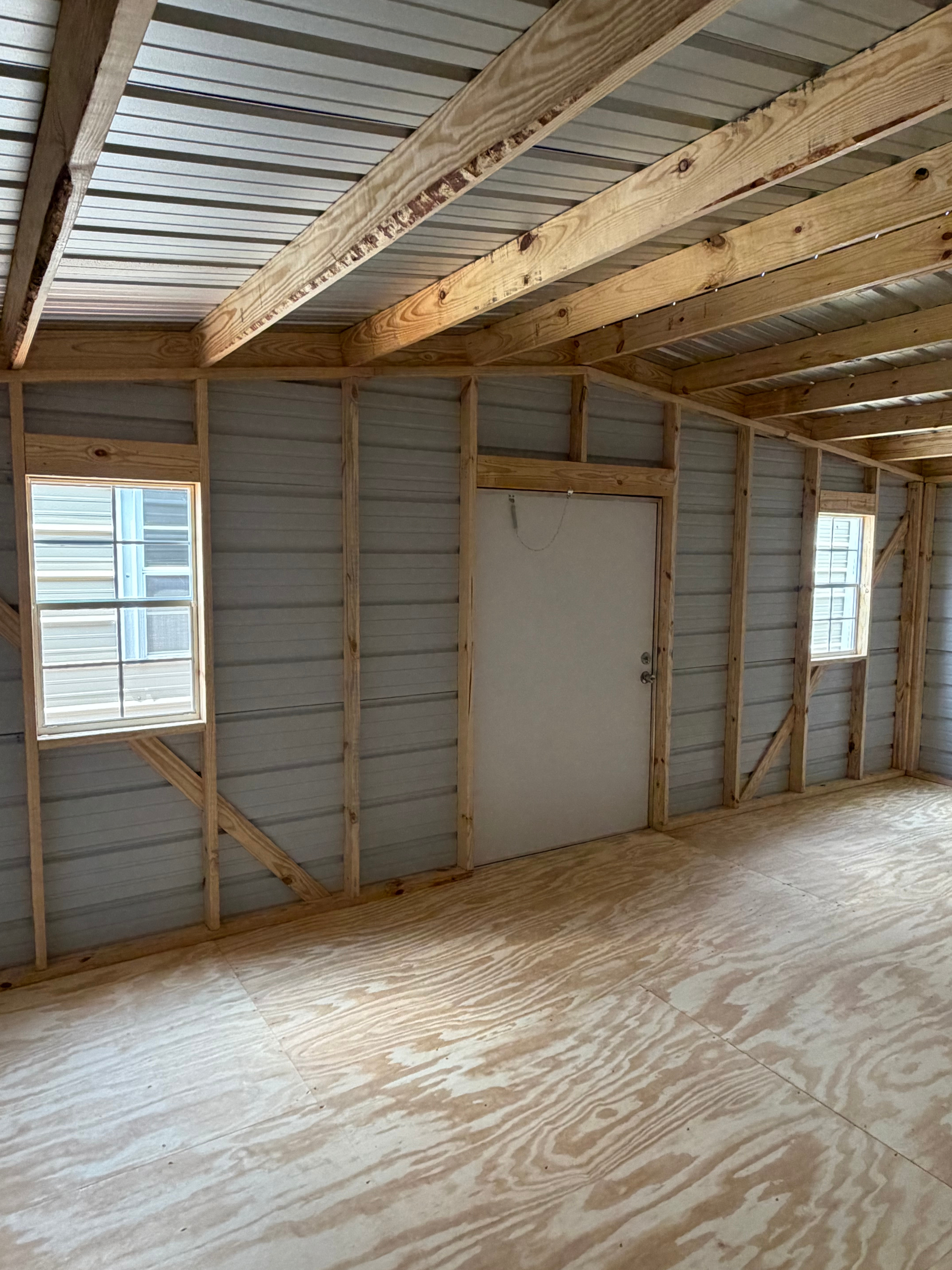 Empty unfinished room with exposed wall studs, plywood floor, and two windows with a centered door.