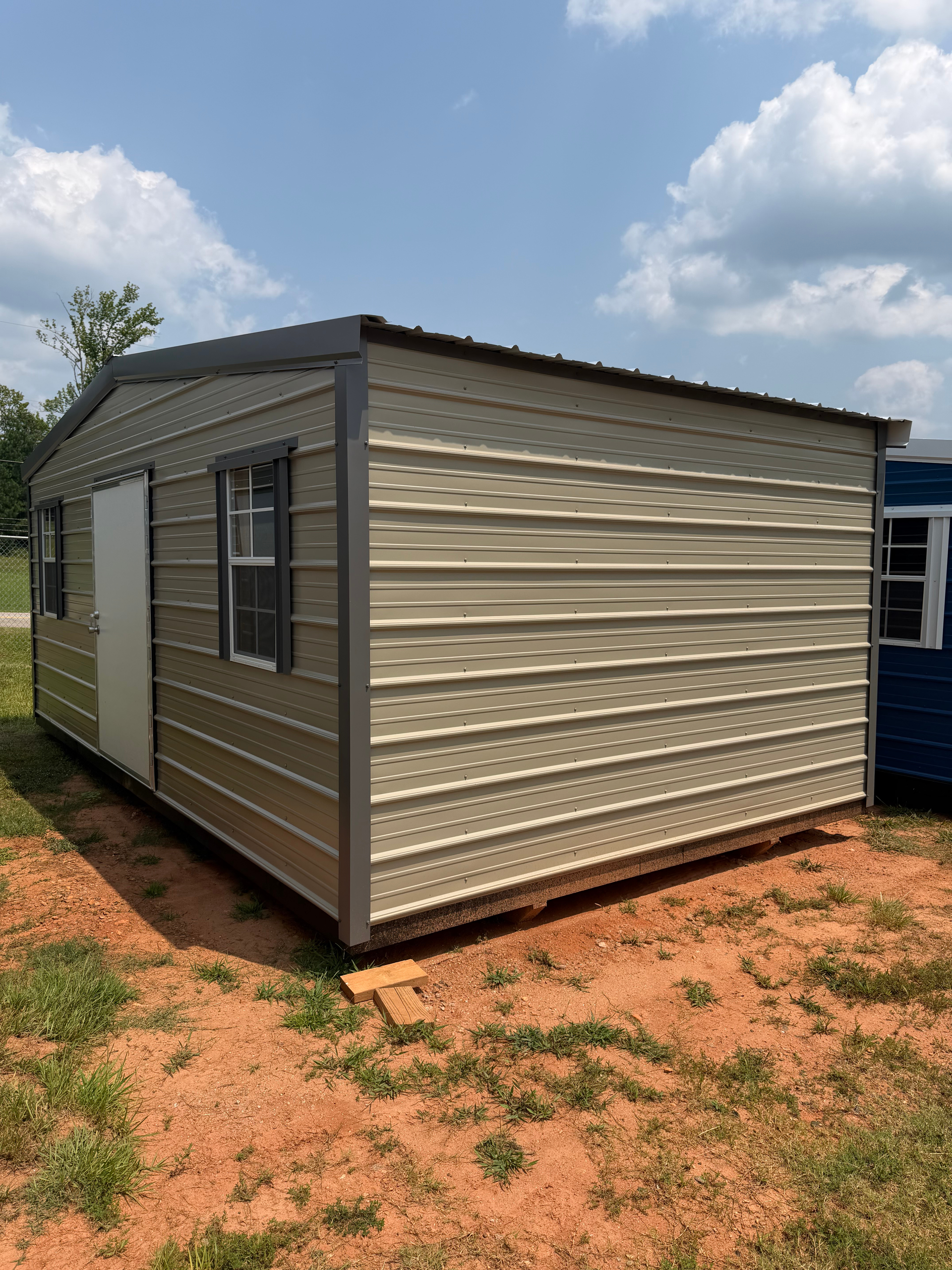 Beige portable shed with brown trim and a window, sitting on red dirt under a cloudy blue sky