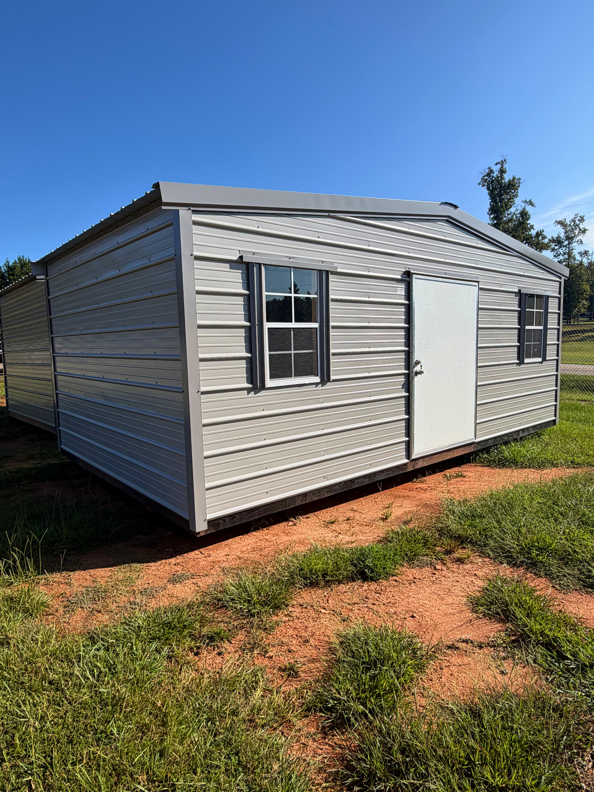 Small gray metal shed with white door and windows on a sunny grassy lot