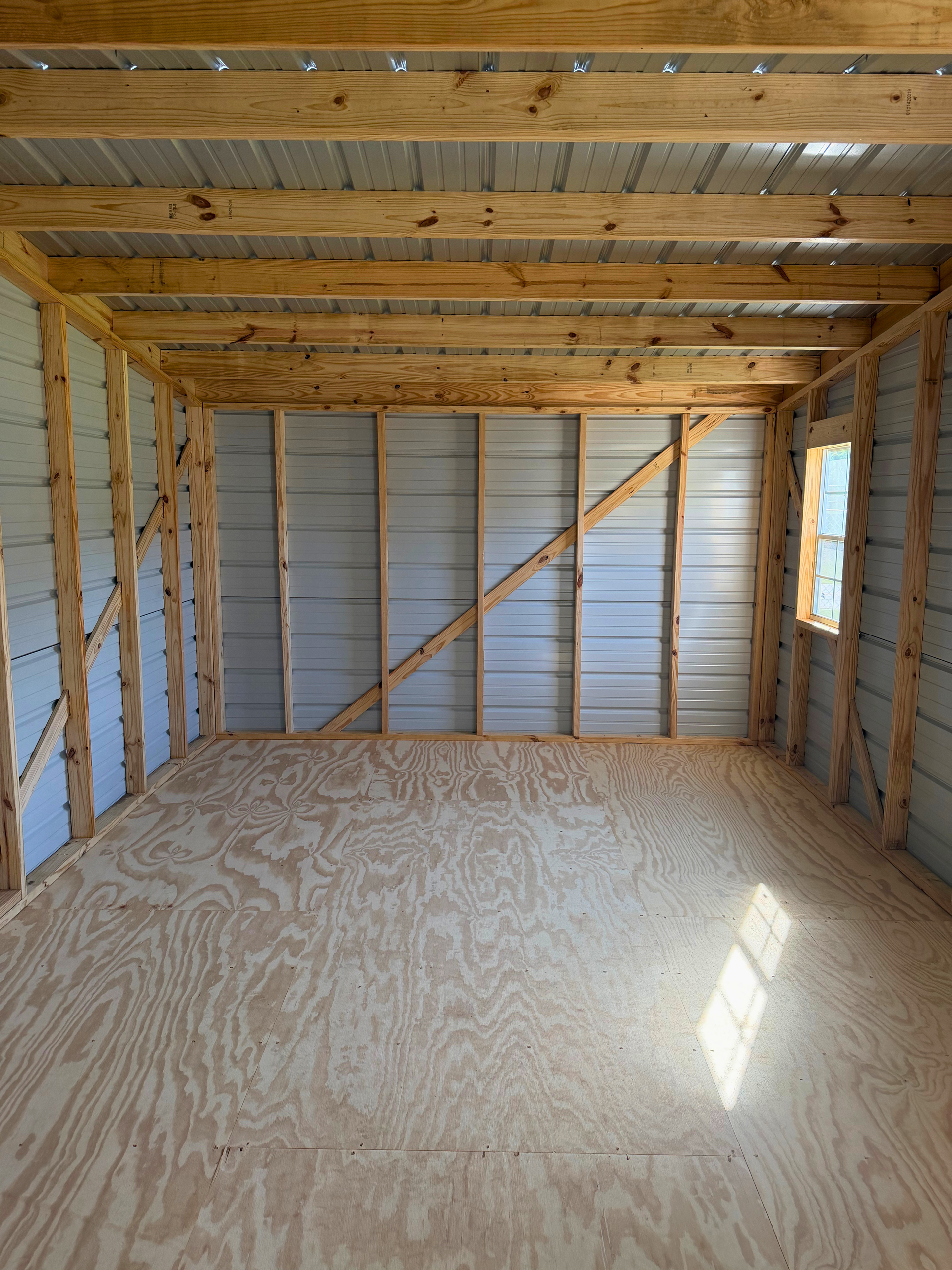 Empty wooden-framed shed interior with plywood floor and a small window on the right.