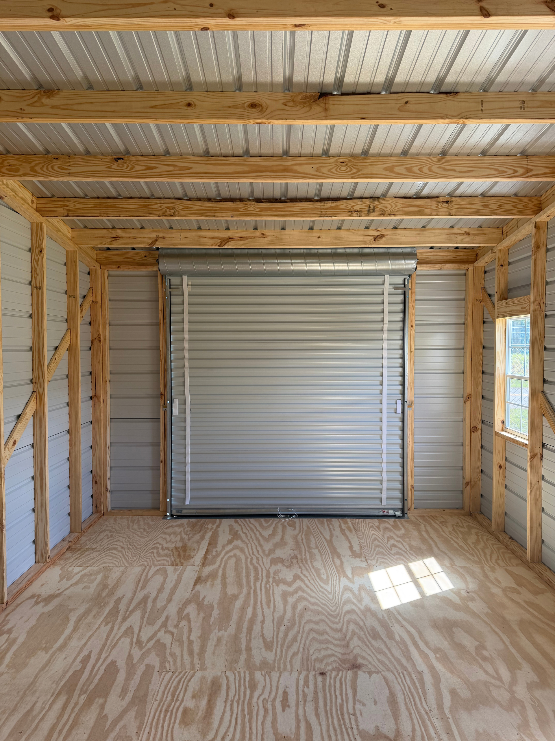 Interior of a wooden-framed shed with a closed white roll-up garage door and plywood floor.
