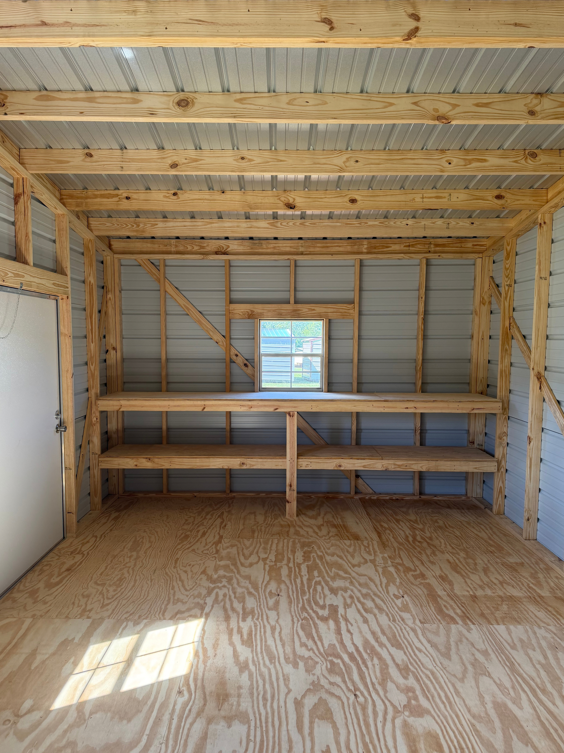 Empty unfinished room with exposed wood framing, plywood floor, shelves, and a small window.
