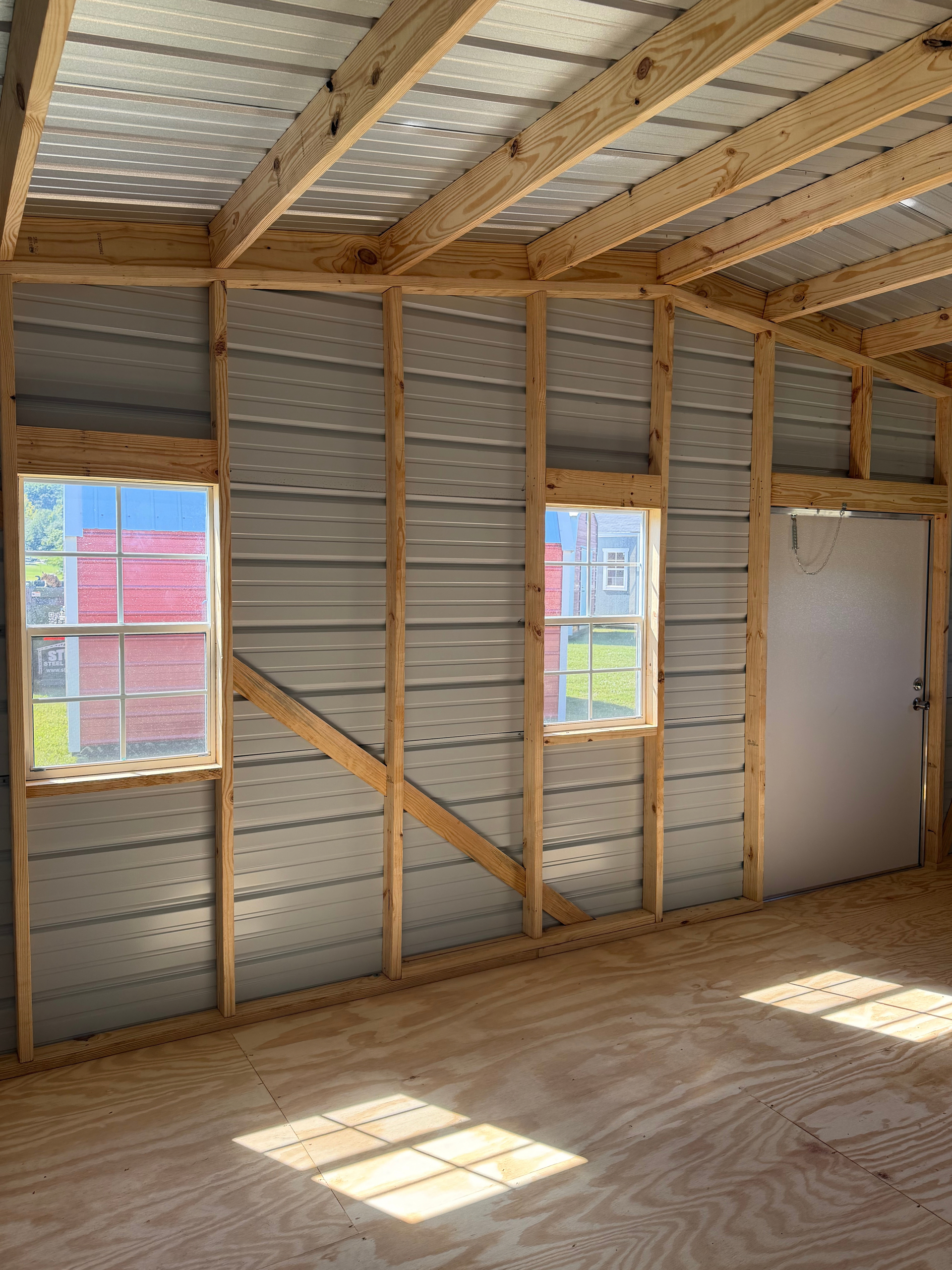 Interior of an unfinished room with exposed framing, two windows, a white door, and sunlight on plywood floor