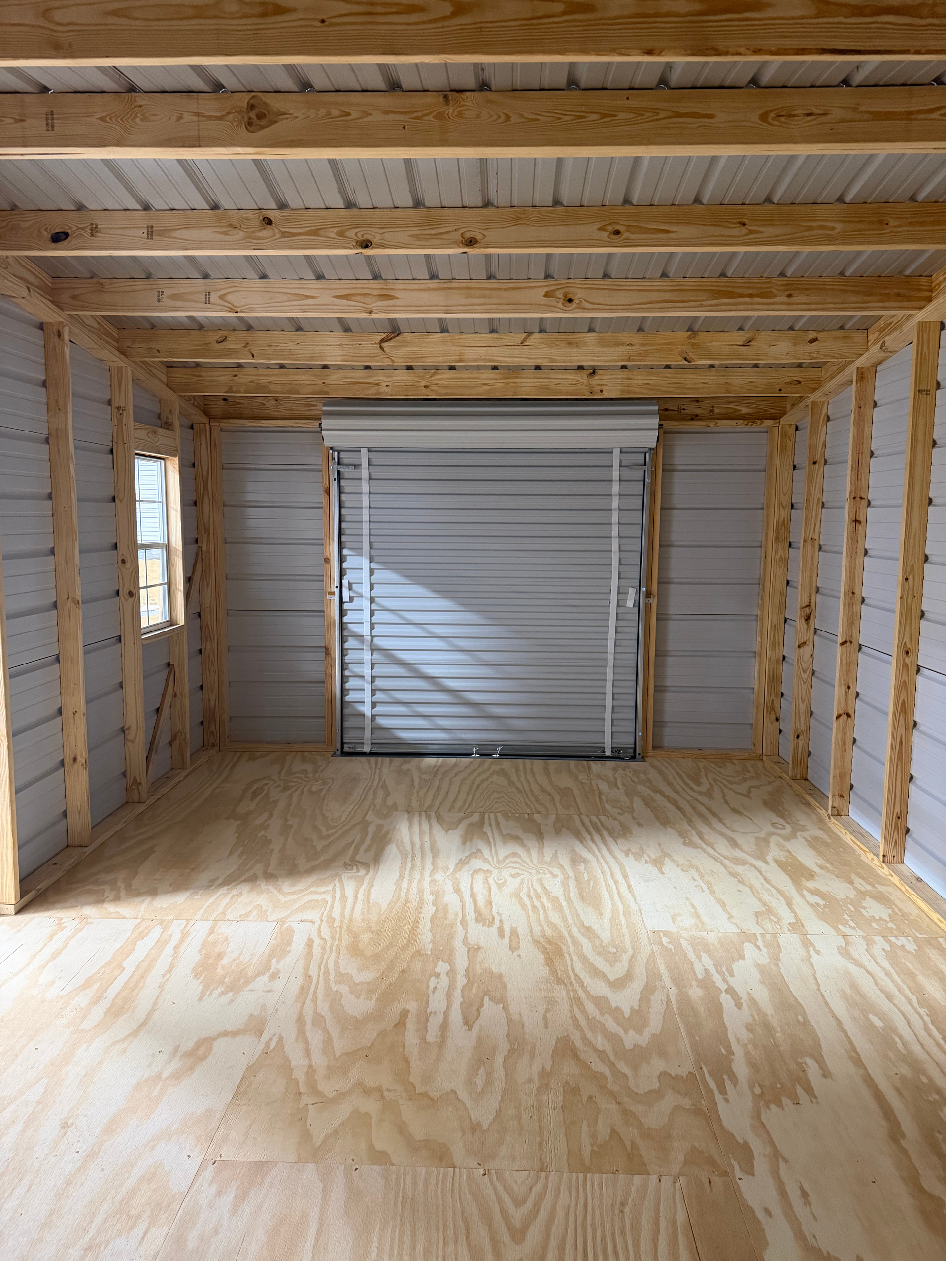 Empty unfinished garage with plywood floor, exposed studs, and a closed roll-up door at the back.