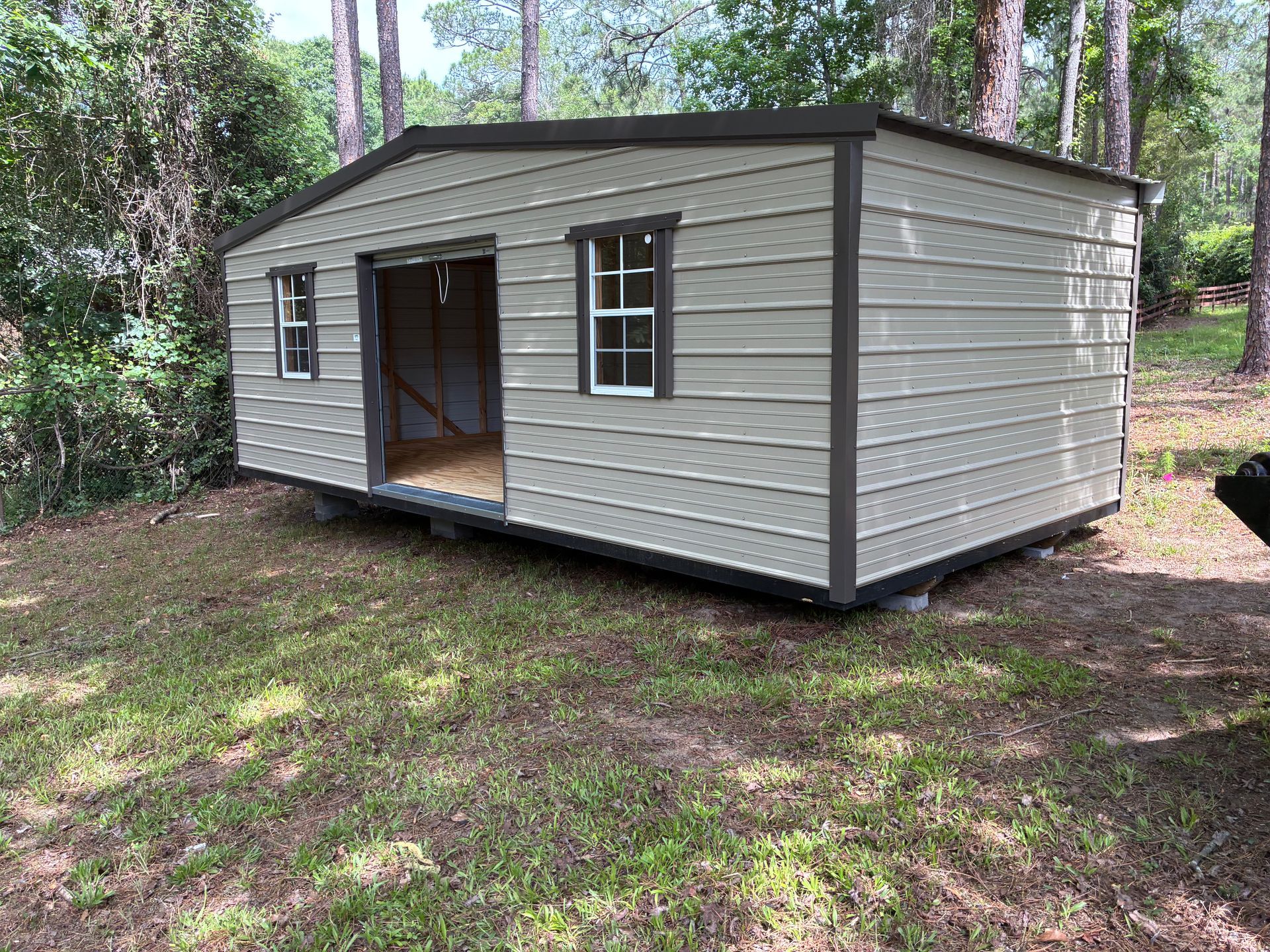 Small beige shed with open doorway and windows in a wooded area.