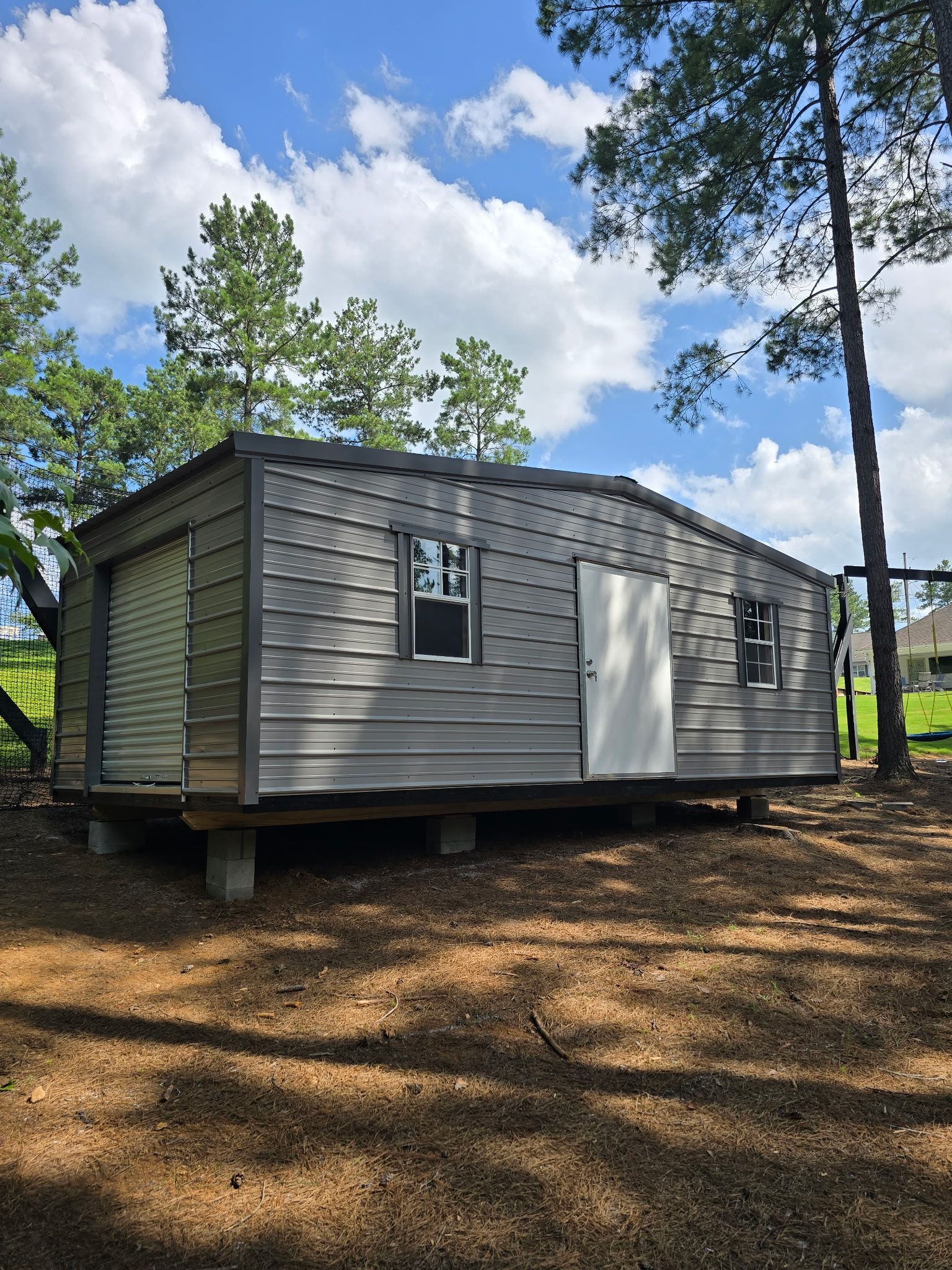 Small gray cabin with white door and windows on a wooden deck in a pine forest under a blue sky.