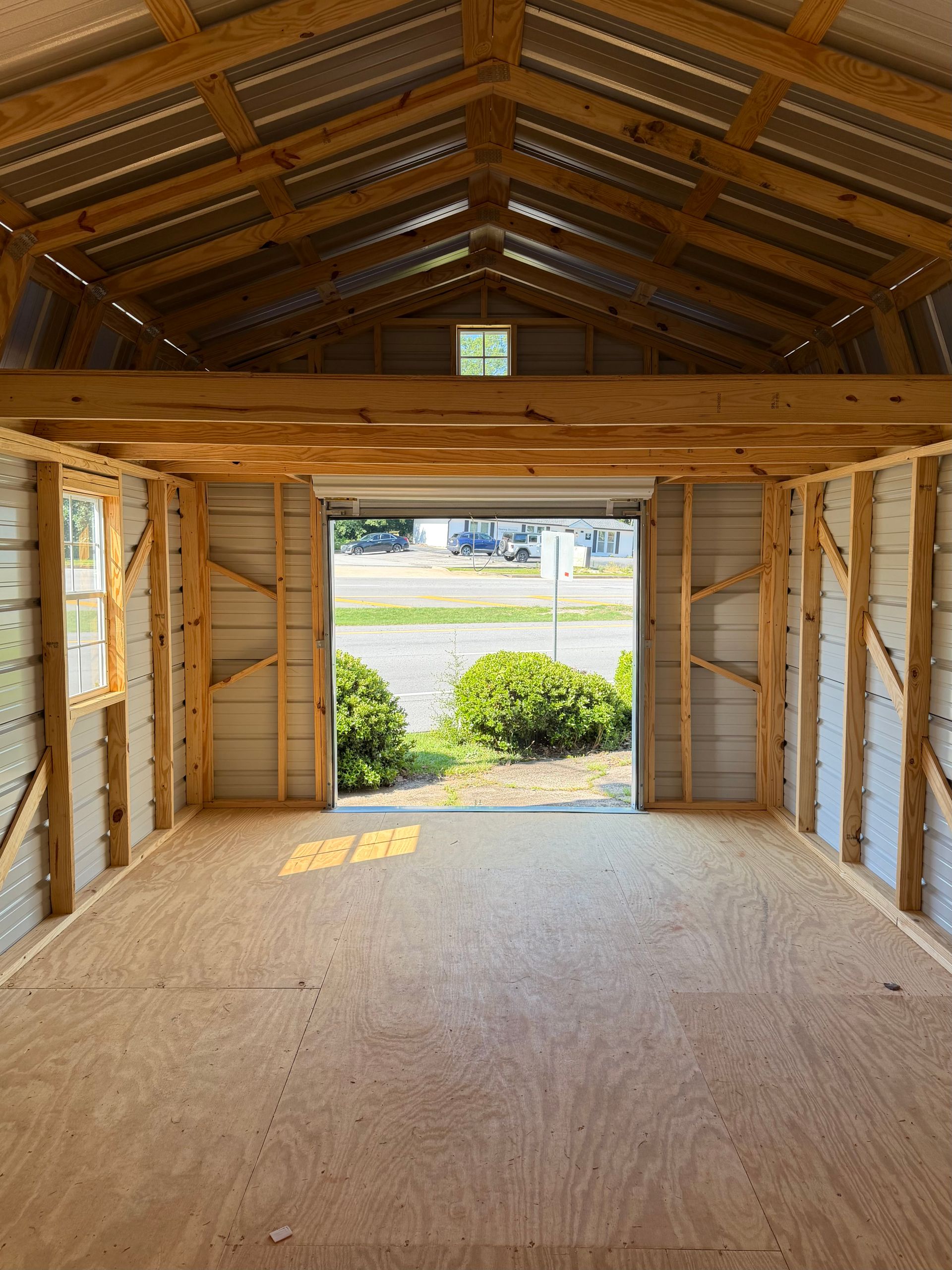 Empty wooden garage interior with open door, sunlight, and view of driveway and bushes outside