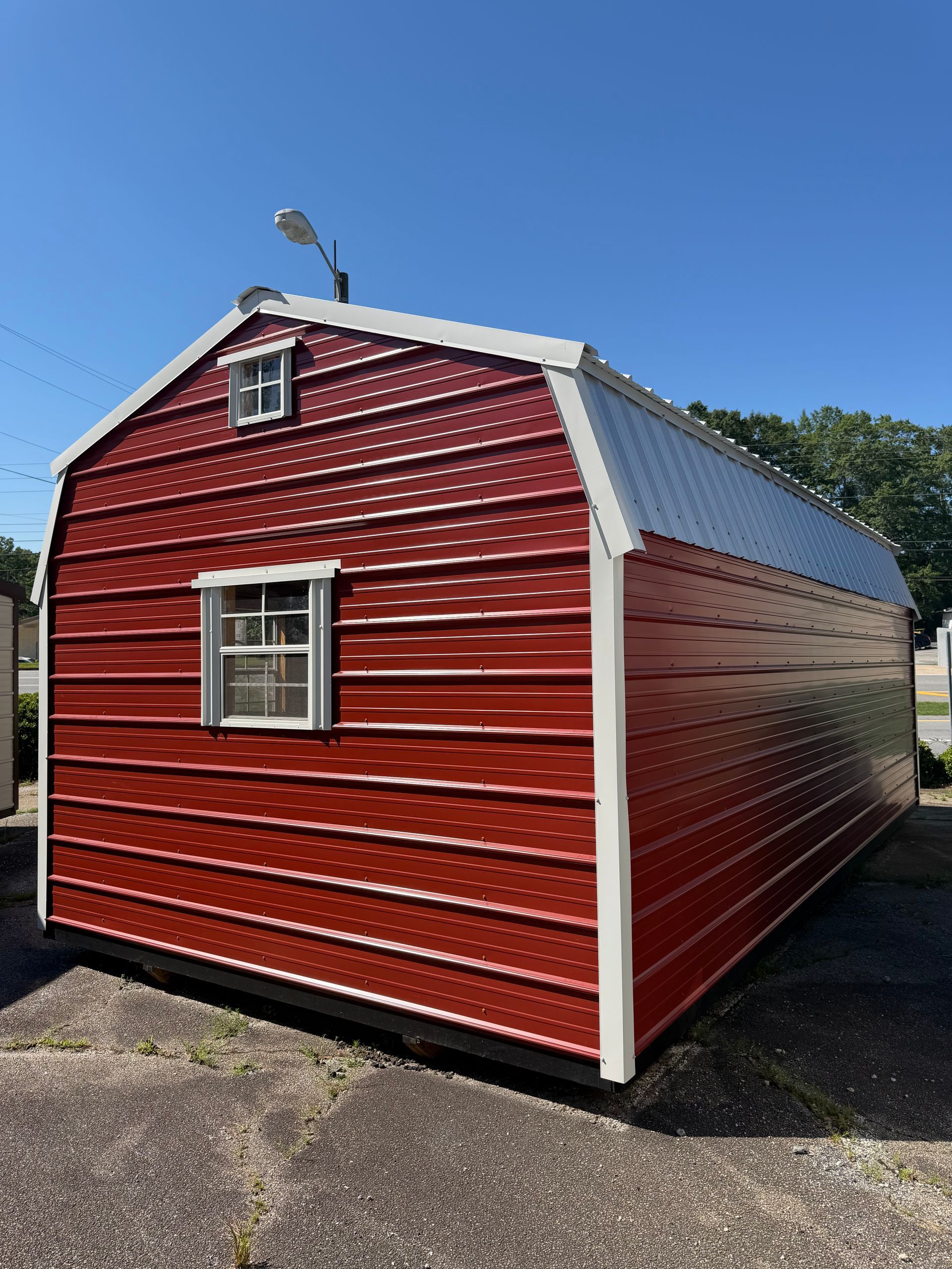 Red metal shed with white trim and a small window on a paved lot under a clear blue sky