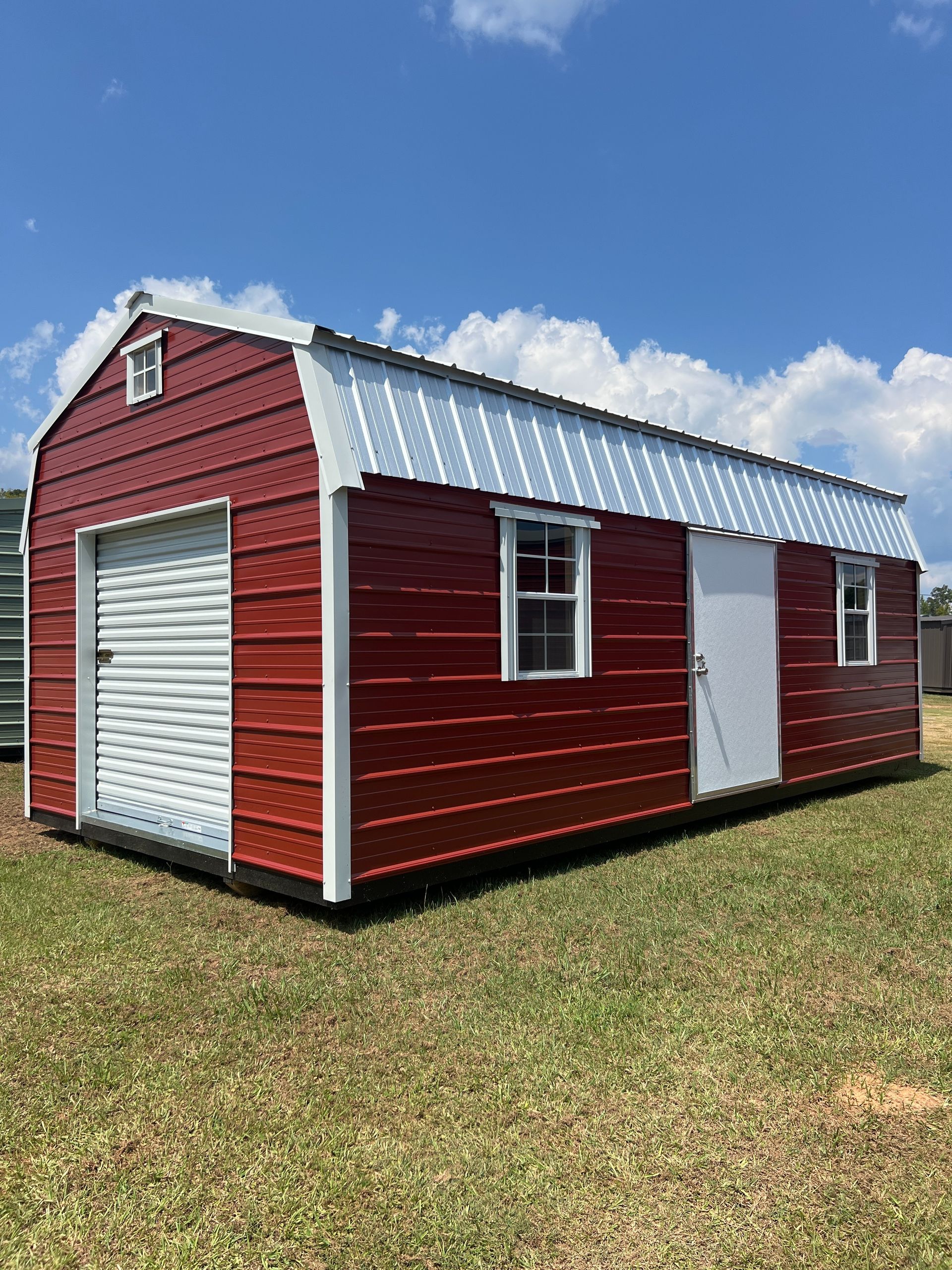 Red shed-style outbuilding with white trim, roll-up door, and a side window on a grassy lawn under blue sky
