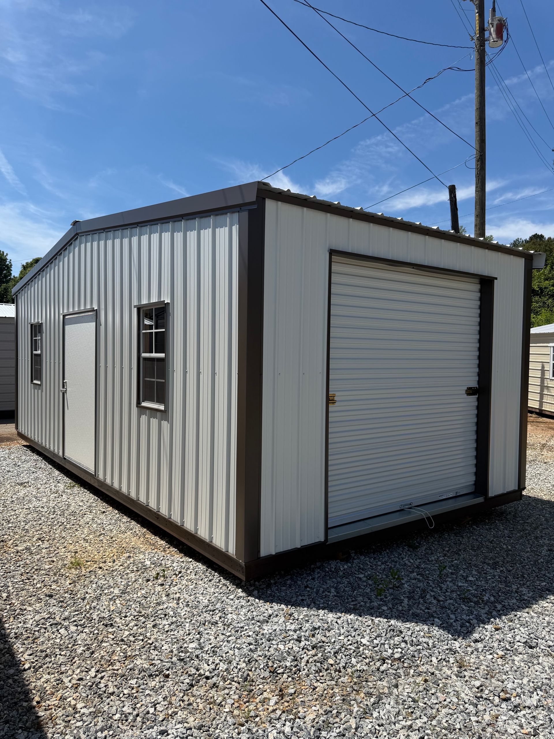 White metal storage shed with a roll-up garage door on a gravel lot under a blue sky