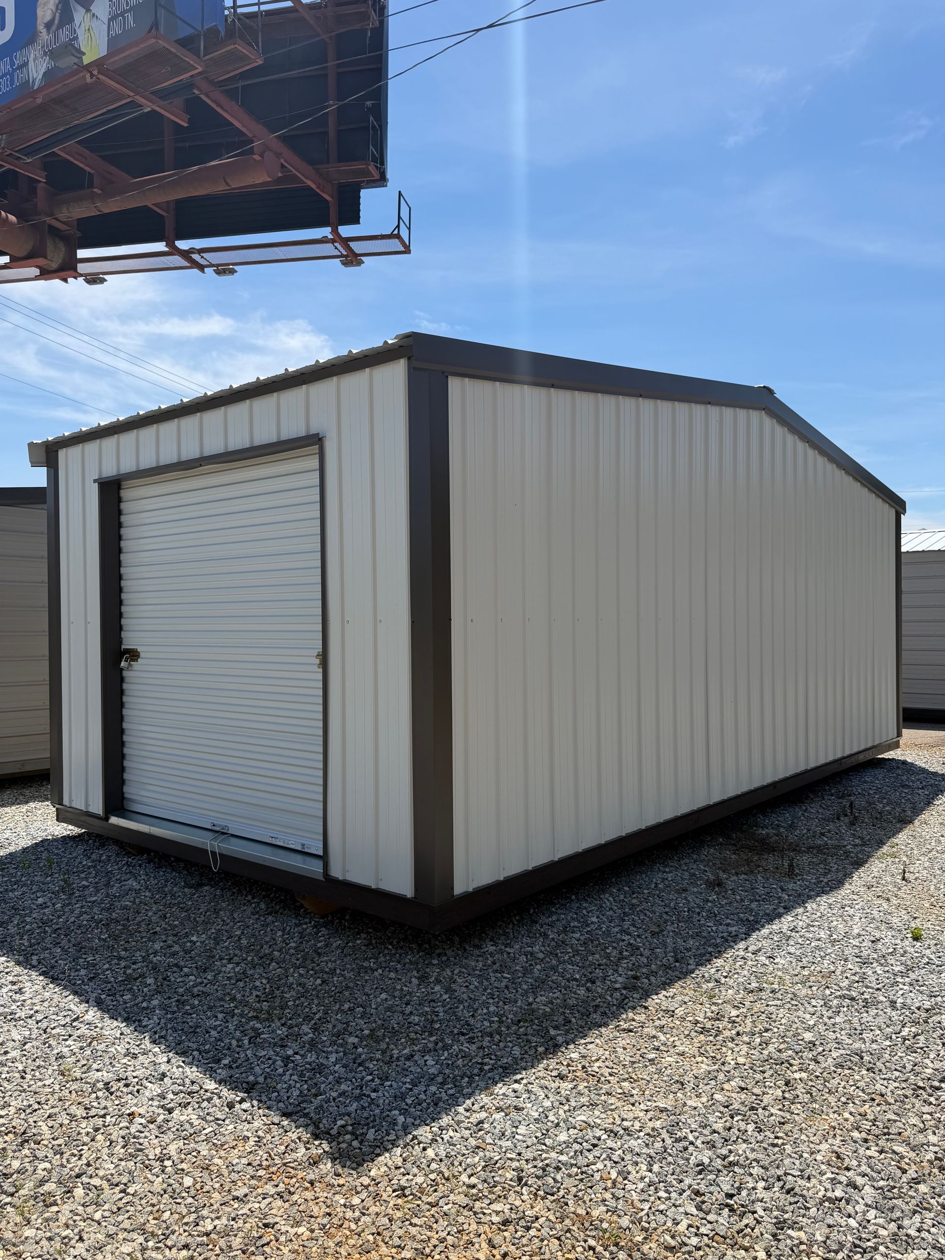 Small beige storage shed with a white roll-up door on a gravel lot under a blue sky.