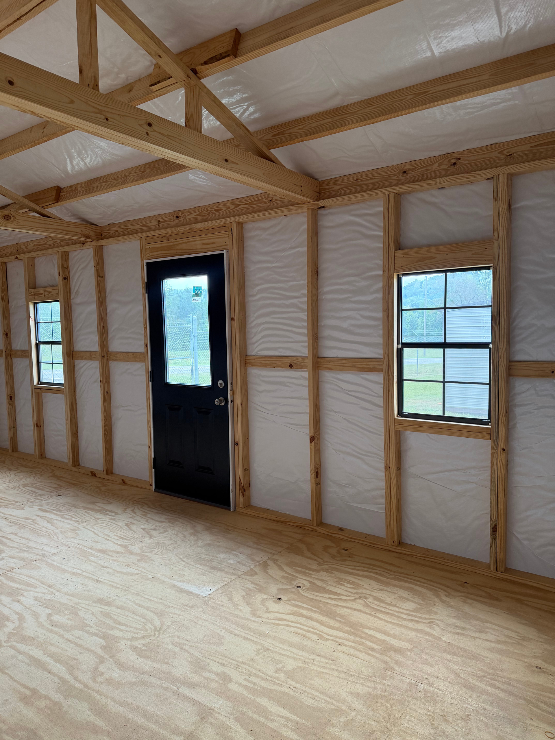 Interior of a newly framed room with exposed wood studs, insulation, plywood subfloor, and a black door.
