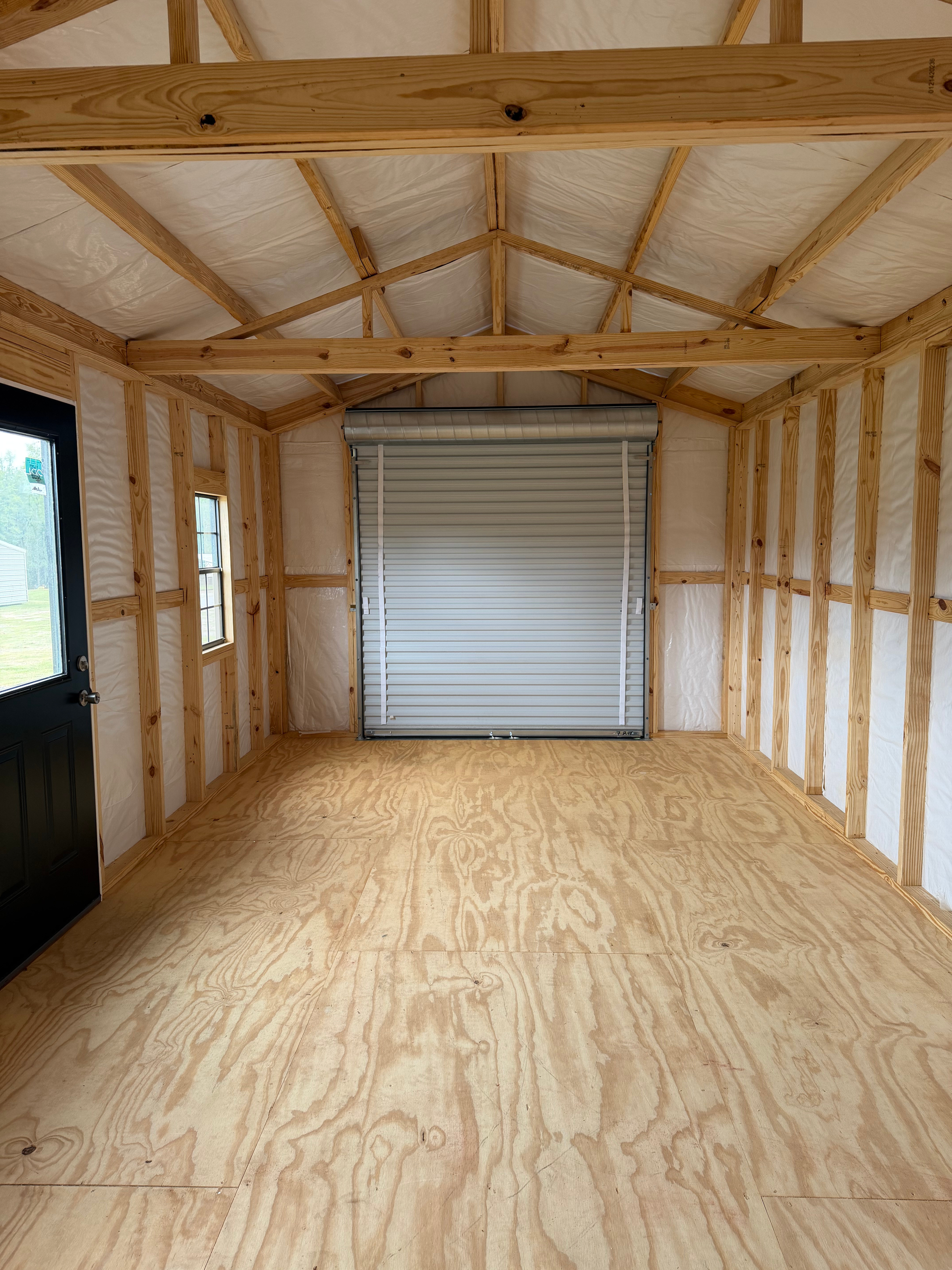 Empty unfinished shed interior with exposed framing, plywood floor, and a closed roll-up door.