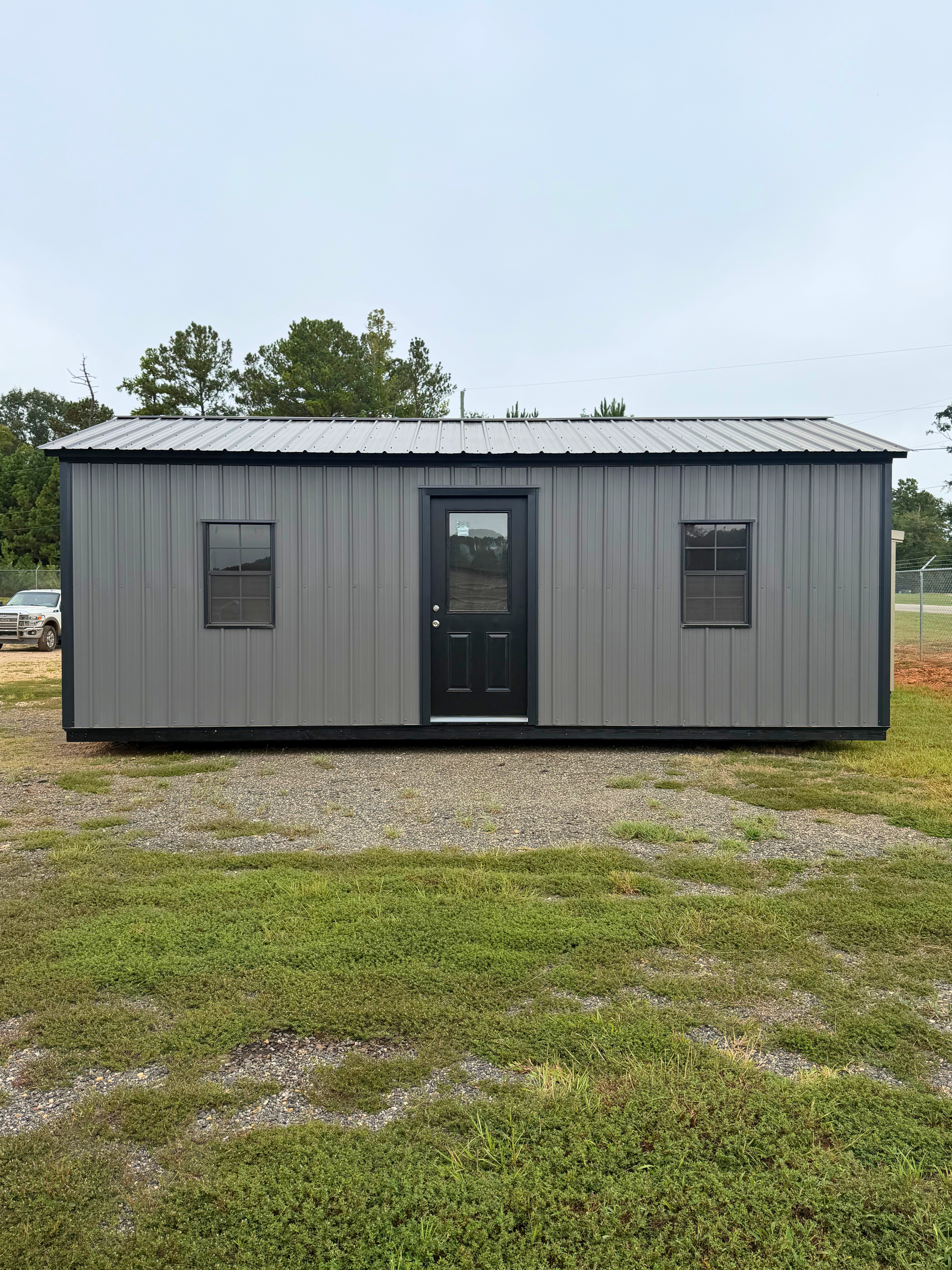 Gray metal tiny house with a black front door and windows, standing on gravel in a grassy field.