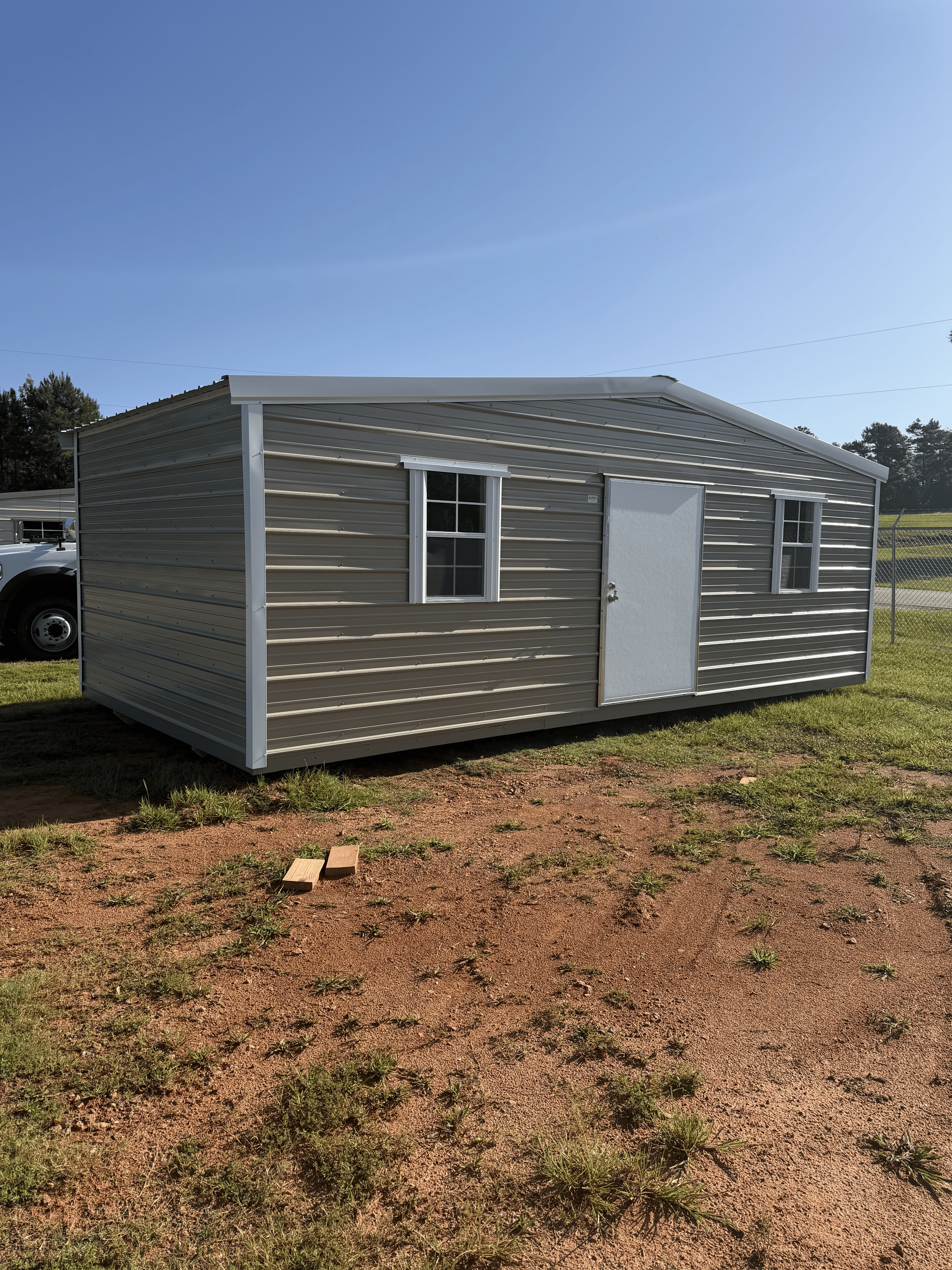 Small gray metal shed with a white door and window, sitting on bare ground under a clear blue sky.