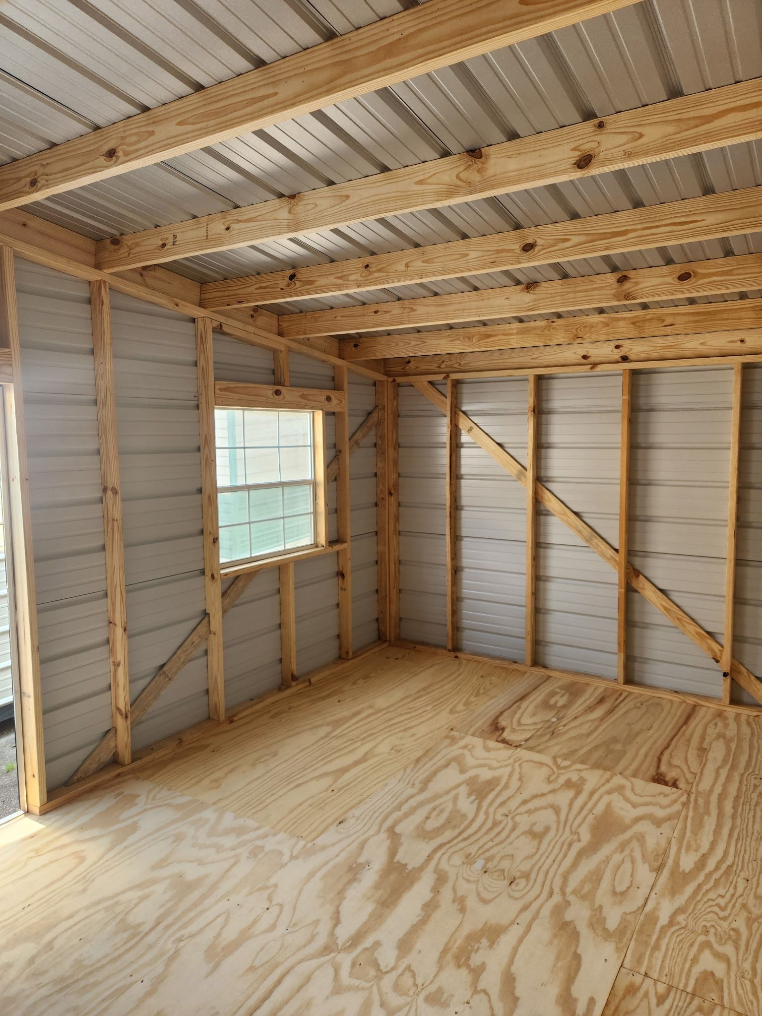 Interior of an unfinished wooden shed with plywood floor, exposed studs, and a small window.