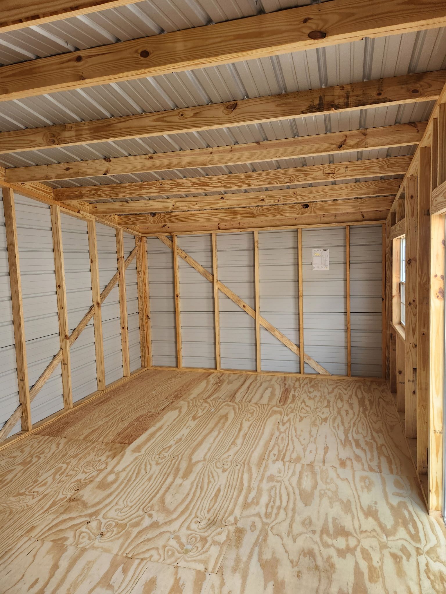 Empty unfinished room with exposed wood framing, plywood floor, and corrugated metal walls and ceiling.
