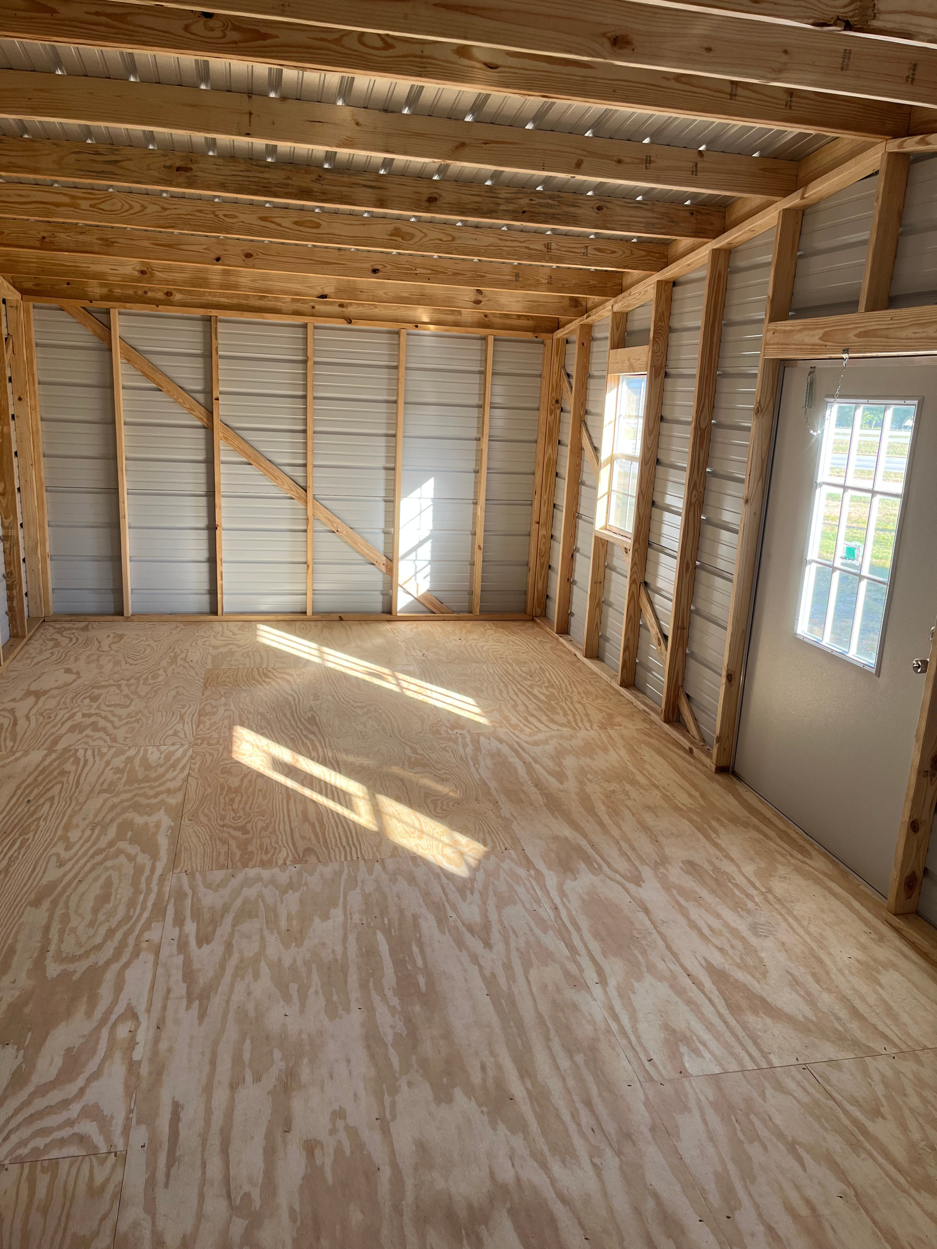 Empty unfinished room with plywood floor, exposed framing, and sunlight through a window and door.