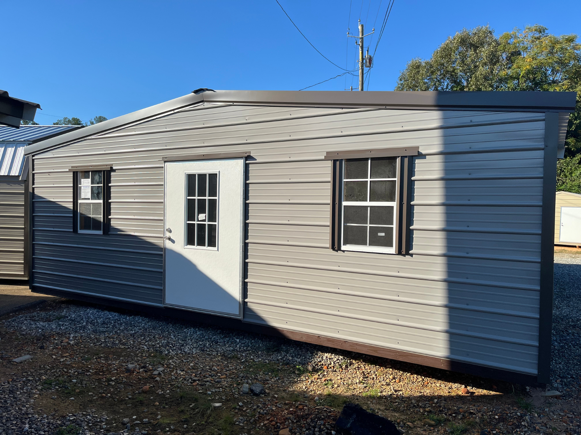 Small beige shed with a white door and dark window shutters on a gravel lot under a blue sky