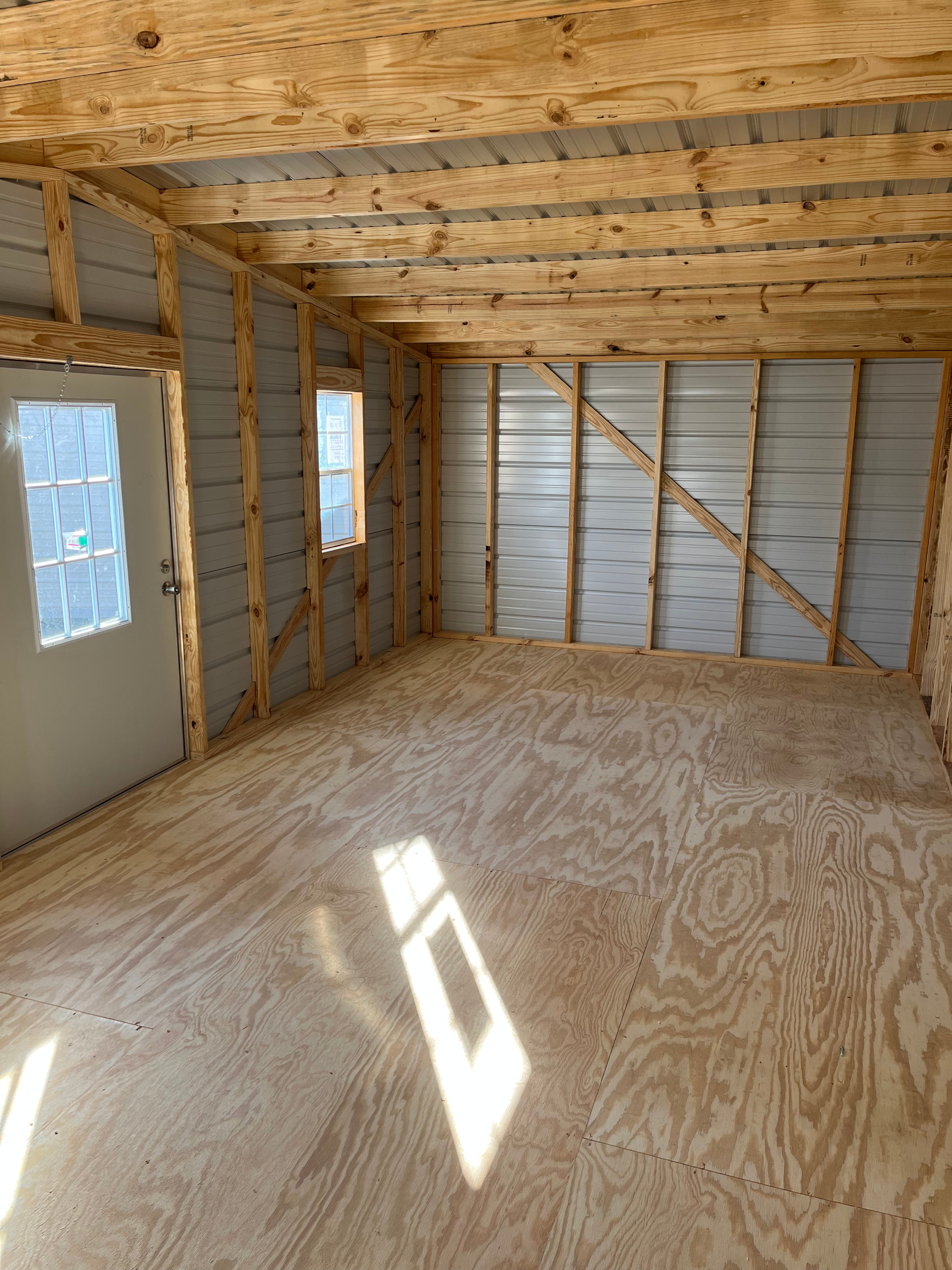 Empty unfinished room with plywood floor, exposed framing, insulation, and light through two windows and a door.