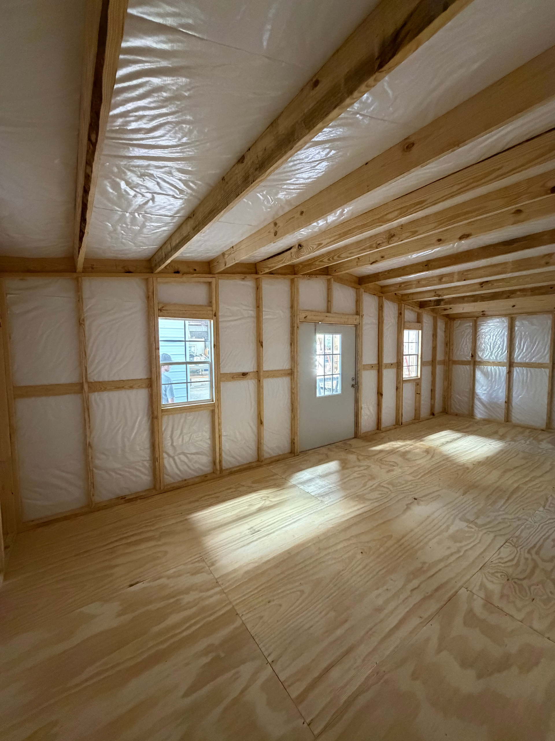 Empty unfinished room with exposed wood framing, insulation, and sunlit windows on a plywood floor