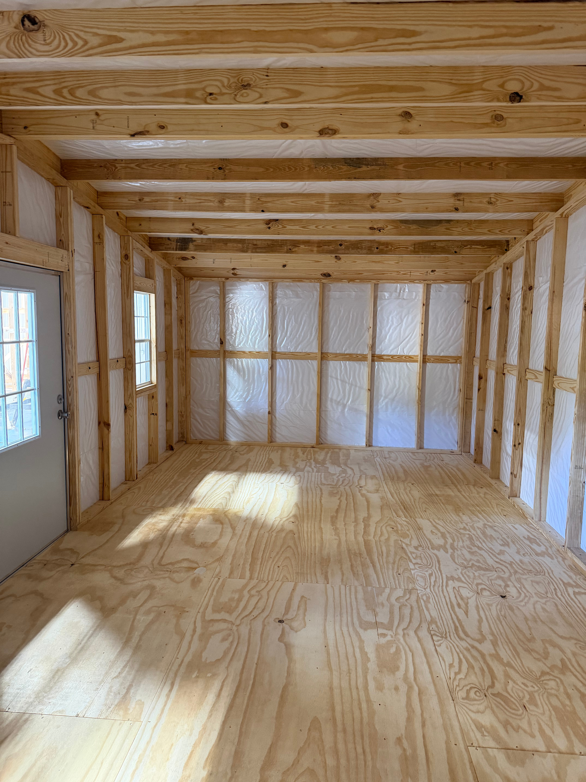 Empty unfinished wooden interior with exposed studs, plywood floor, and small windows.