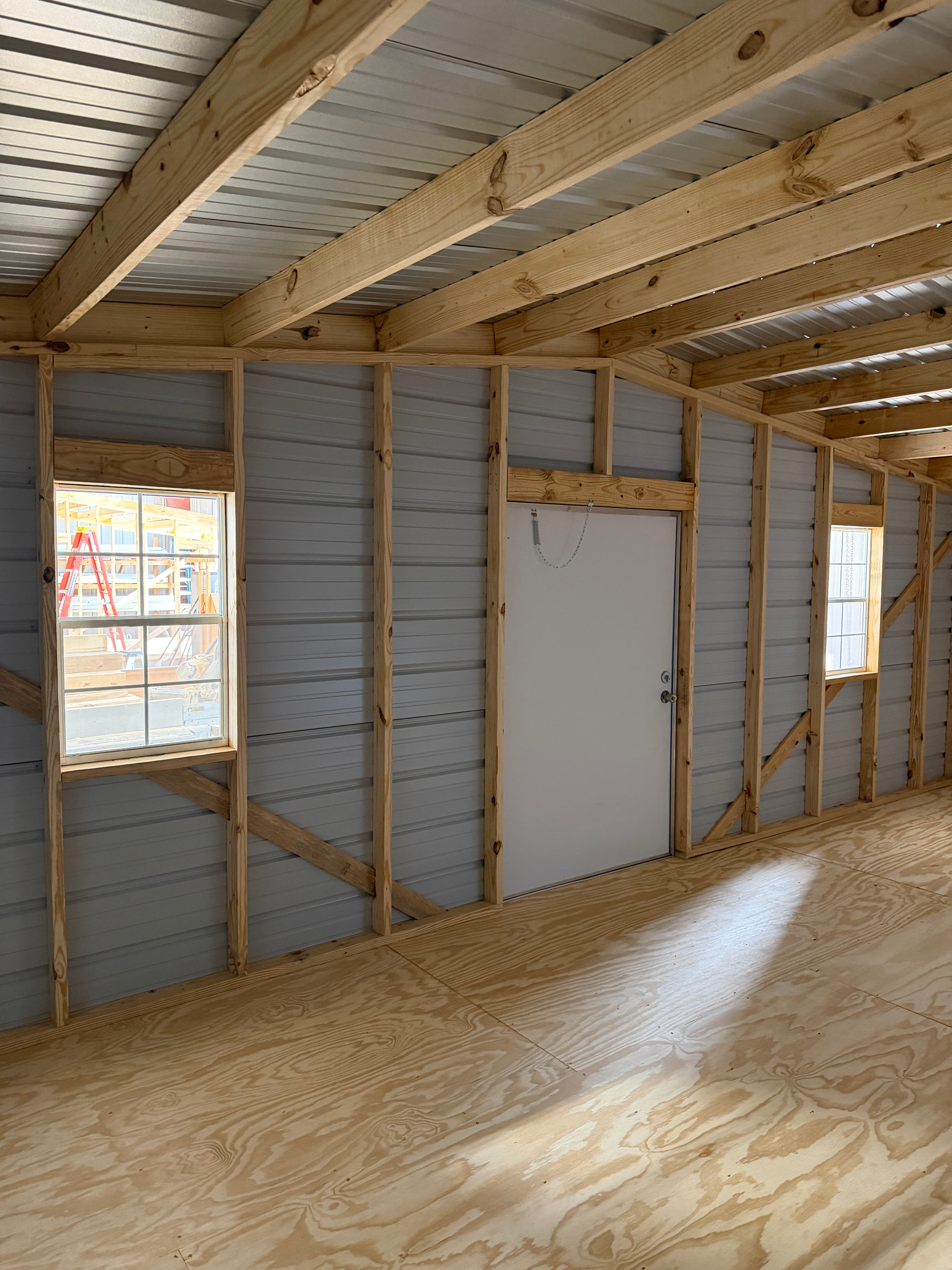 Interior of an unfinished room with exposed wood framing, gray sheathing, windows, and a white door.