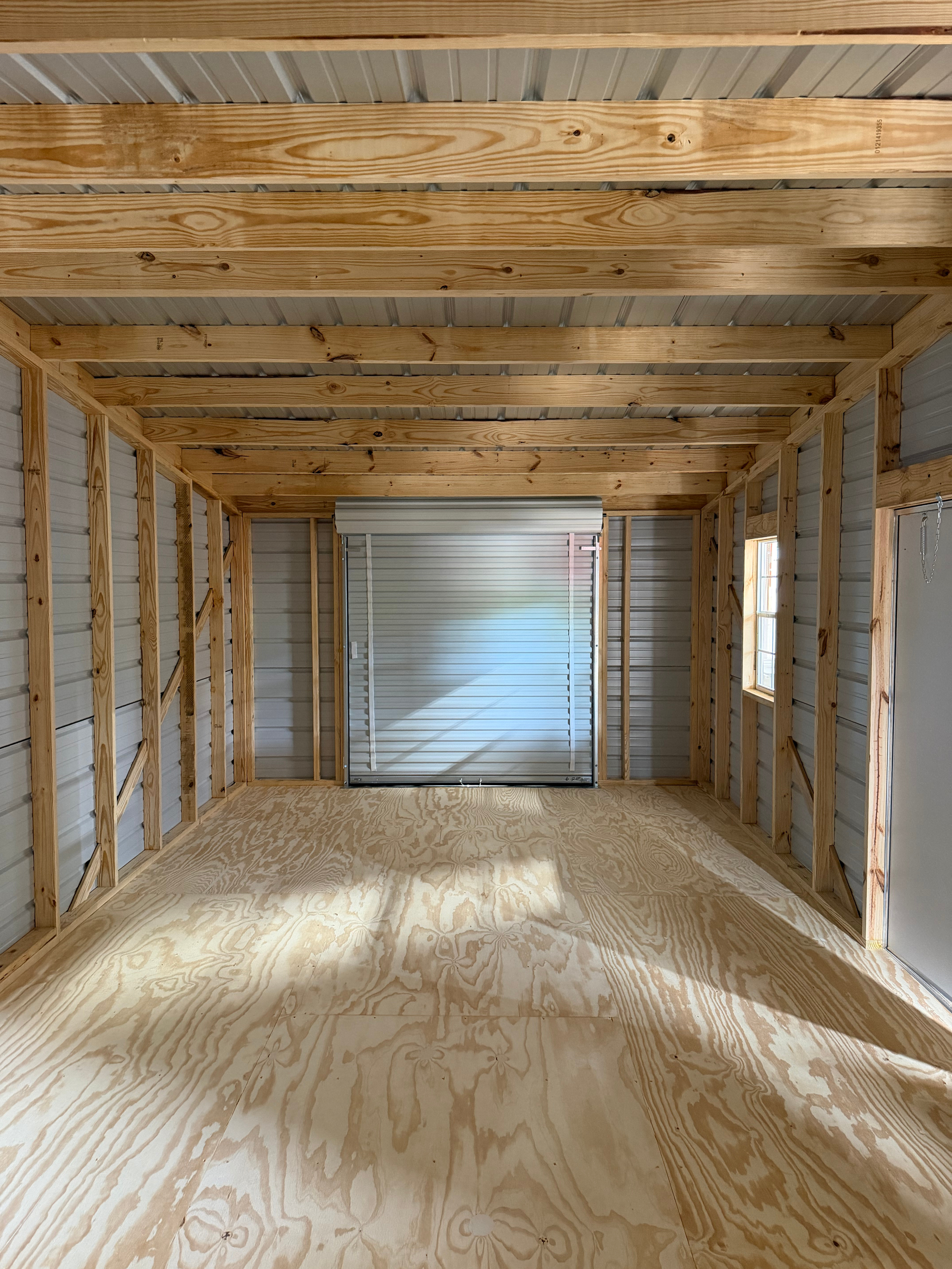 Empty unfinished room with exposed wood framing, plywood floor, and a large opening at the far end.