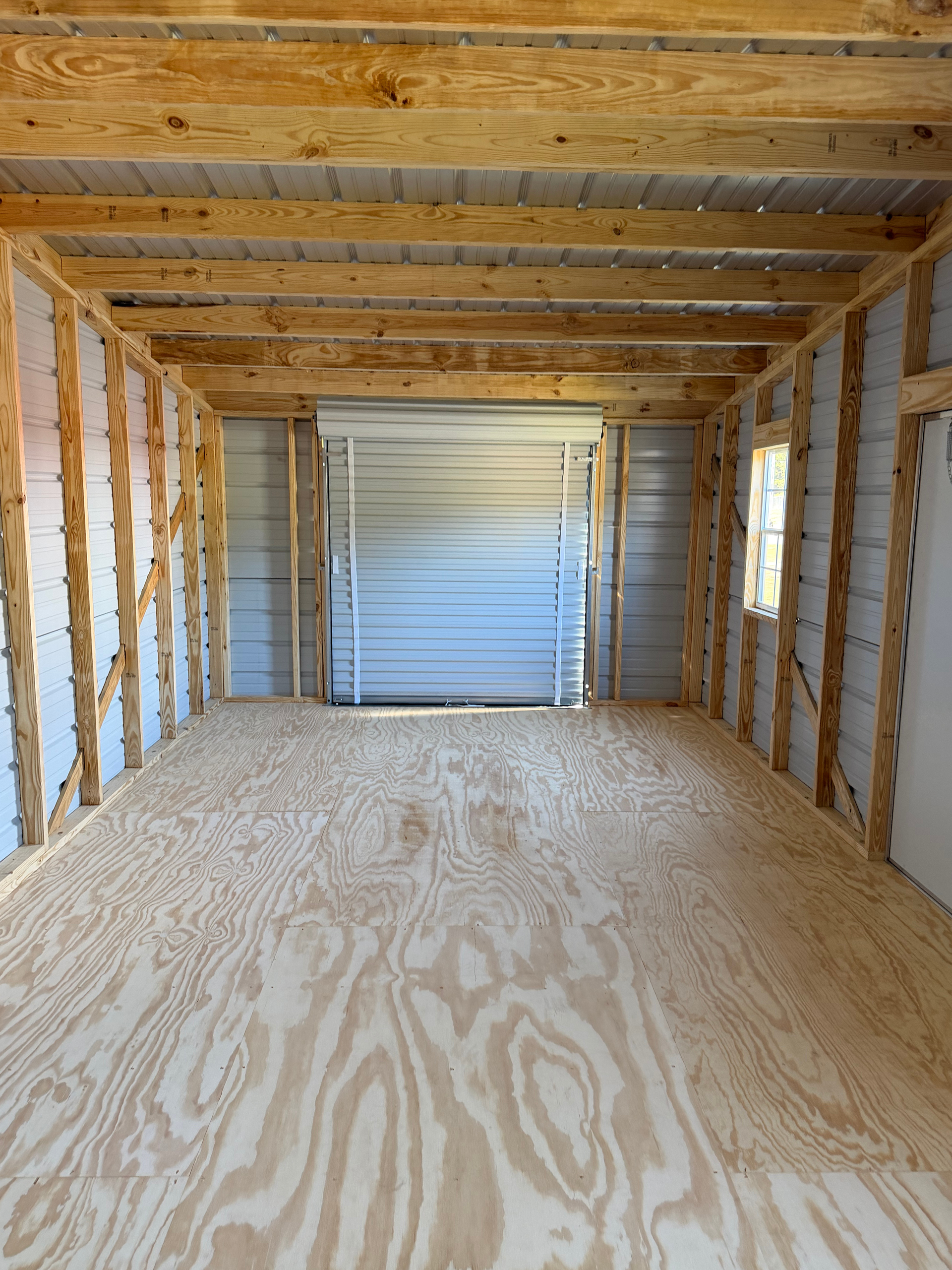 Empty unfinished garage with plywood floor, exposed wooden framing, and a closed metal door at the far end