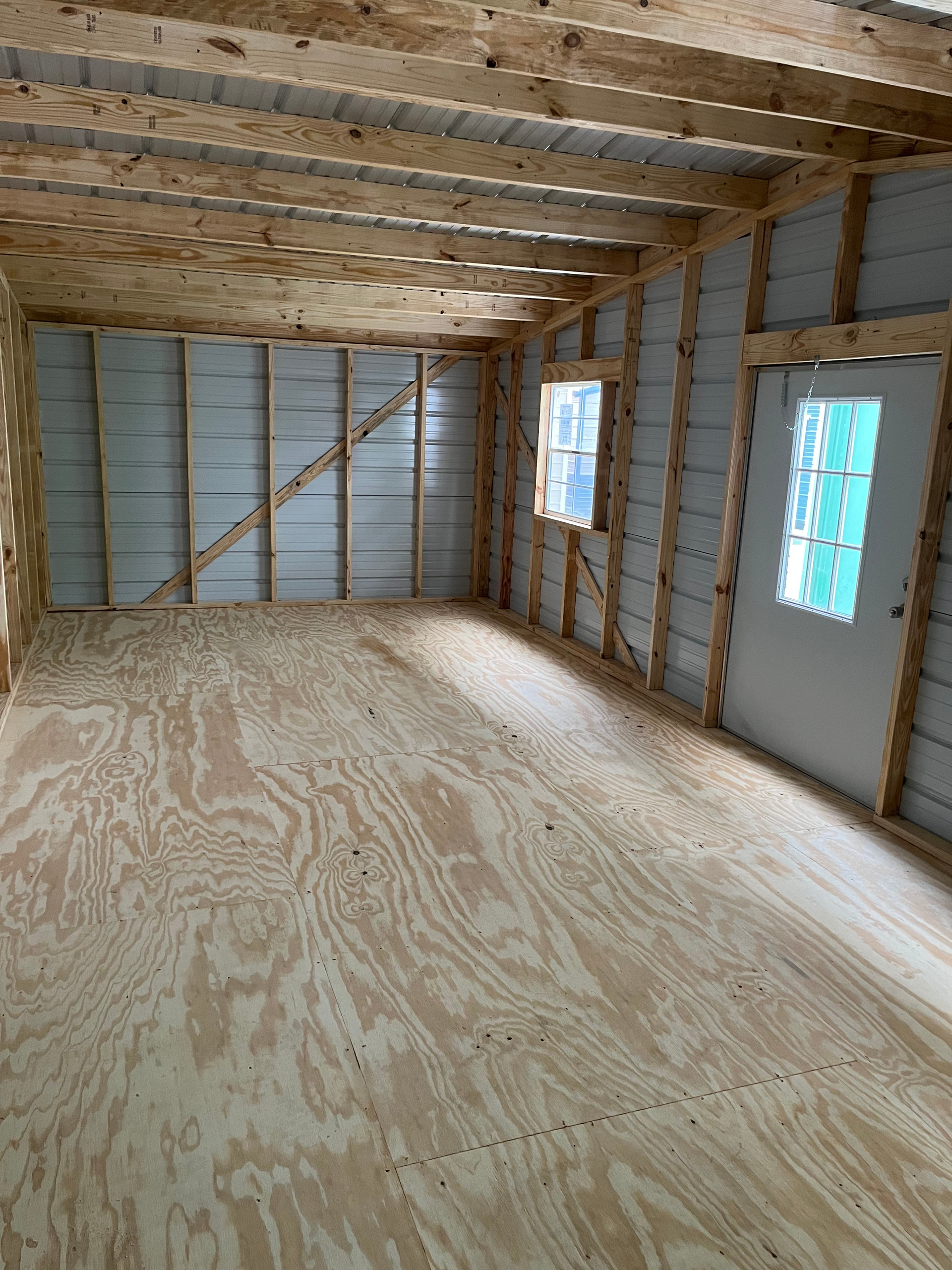 Empty unfinished room with plywood subfloor, exposed studs, and two small windows.