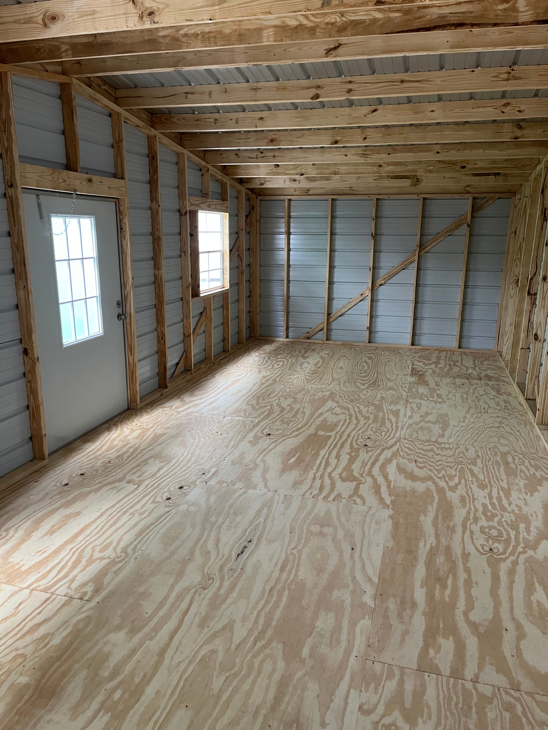 Unfinished room with exposed studs, plywood floor, and two small windows.