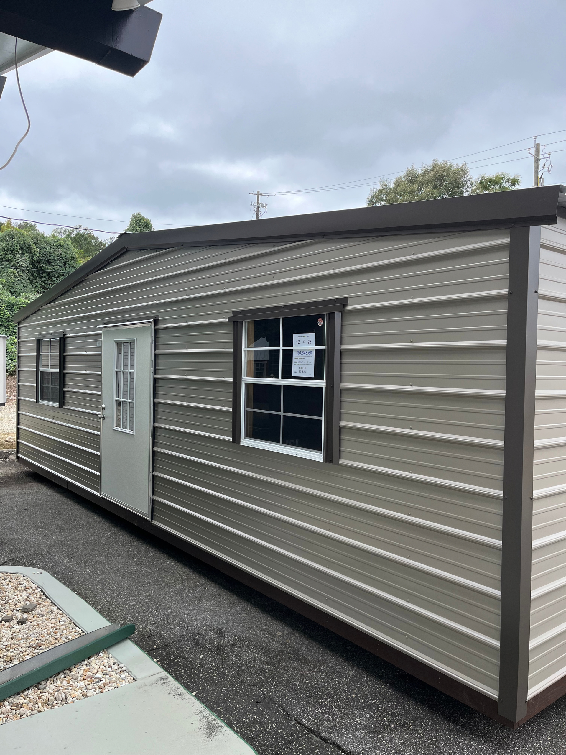 Small beige portable cabin with white door and windows on a gravel lot under cloudy skies