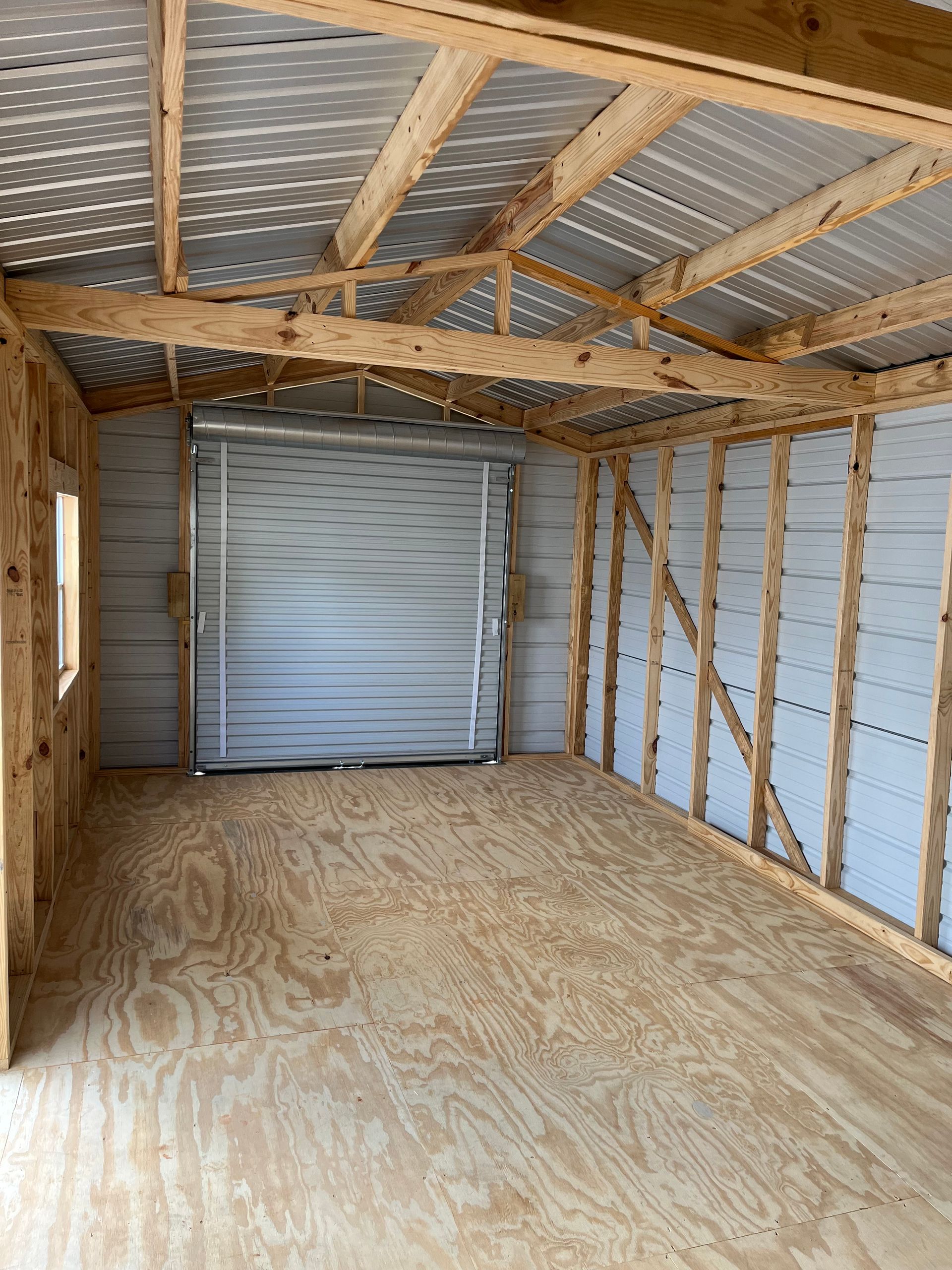Empty unfinished shed with plywood floor, exposed framing, and a closed metal roll-up door.