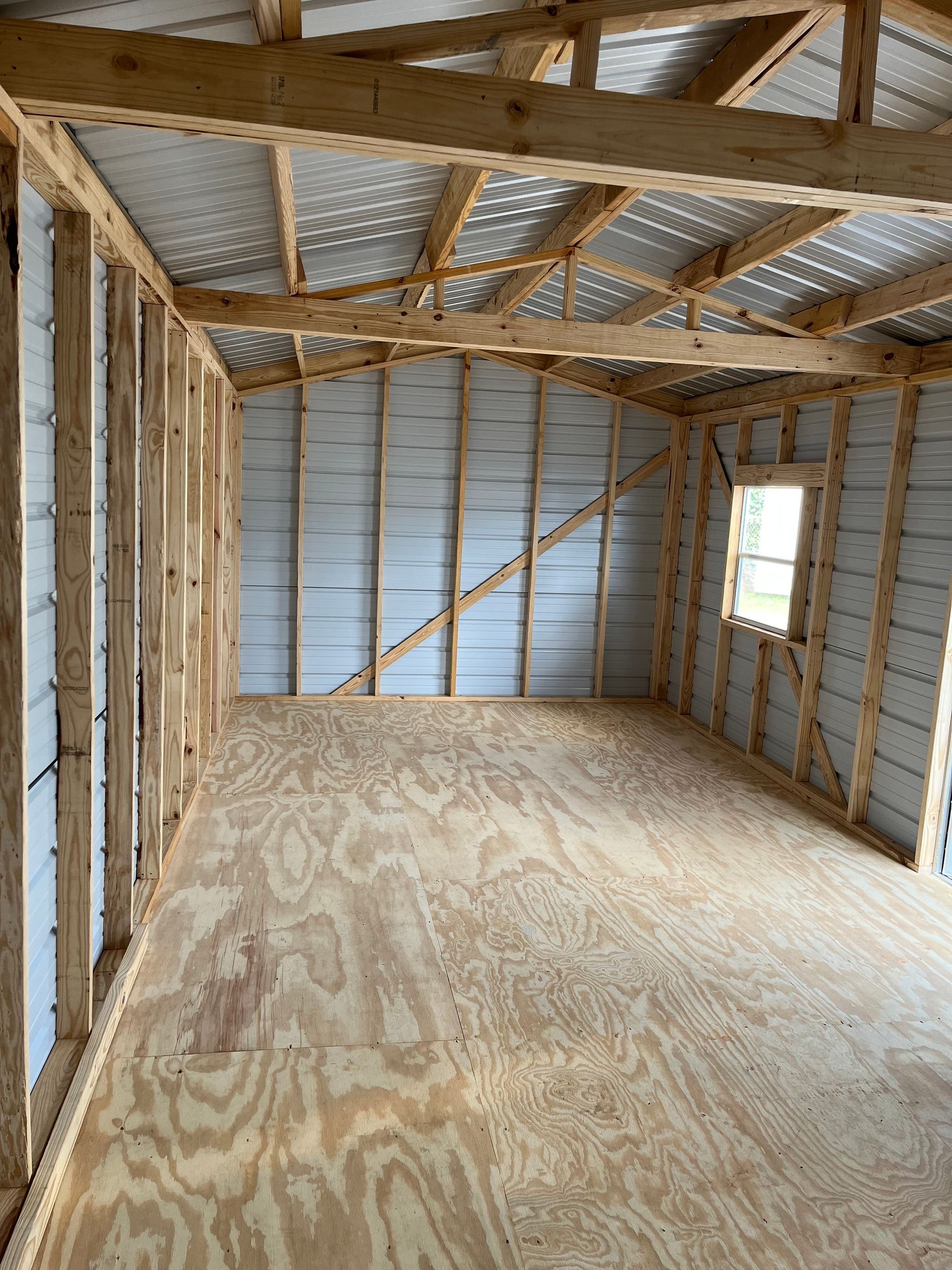 Interior of an unfinished wooden room with exposed framing, plywood floor, and a small window.