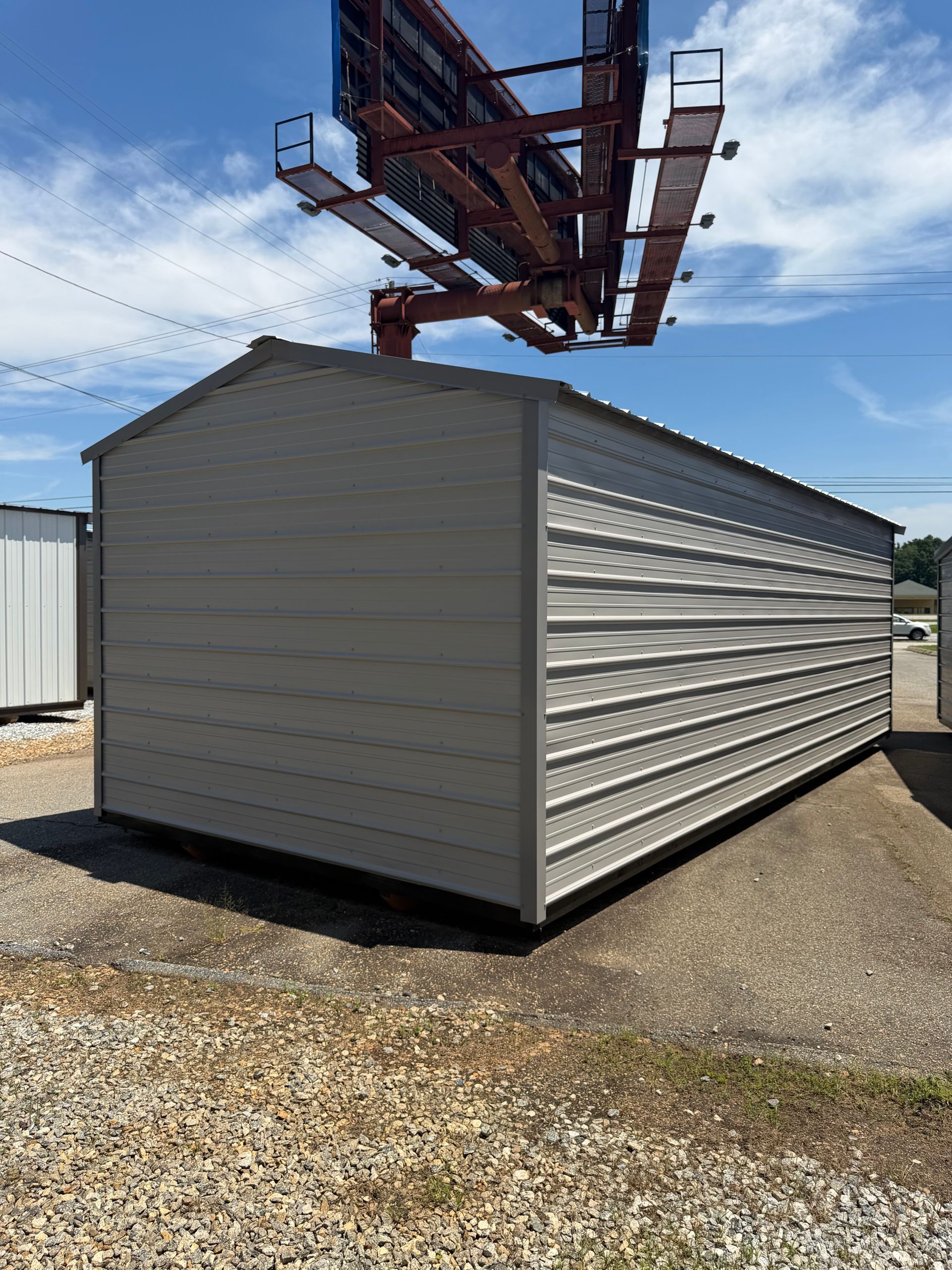 White metal storage shed with a red boom lift overhead on a gravel lot under a blue sky