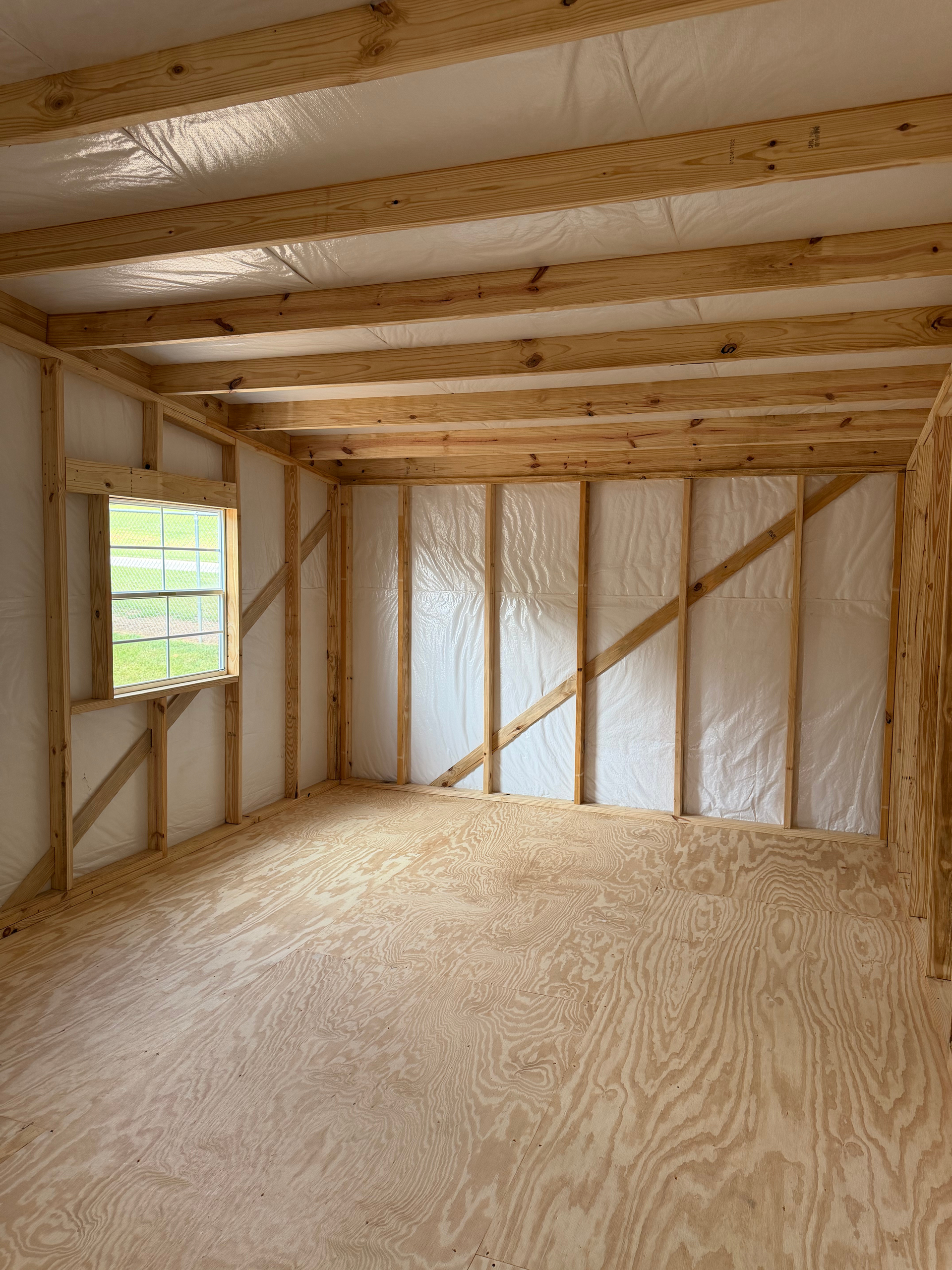 Empty framed room under construction with plywood floor, exposed studs, ceiling joists, and a small window.