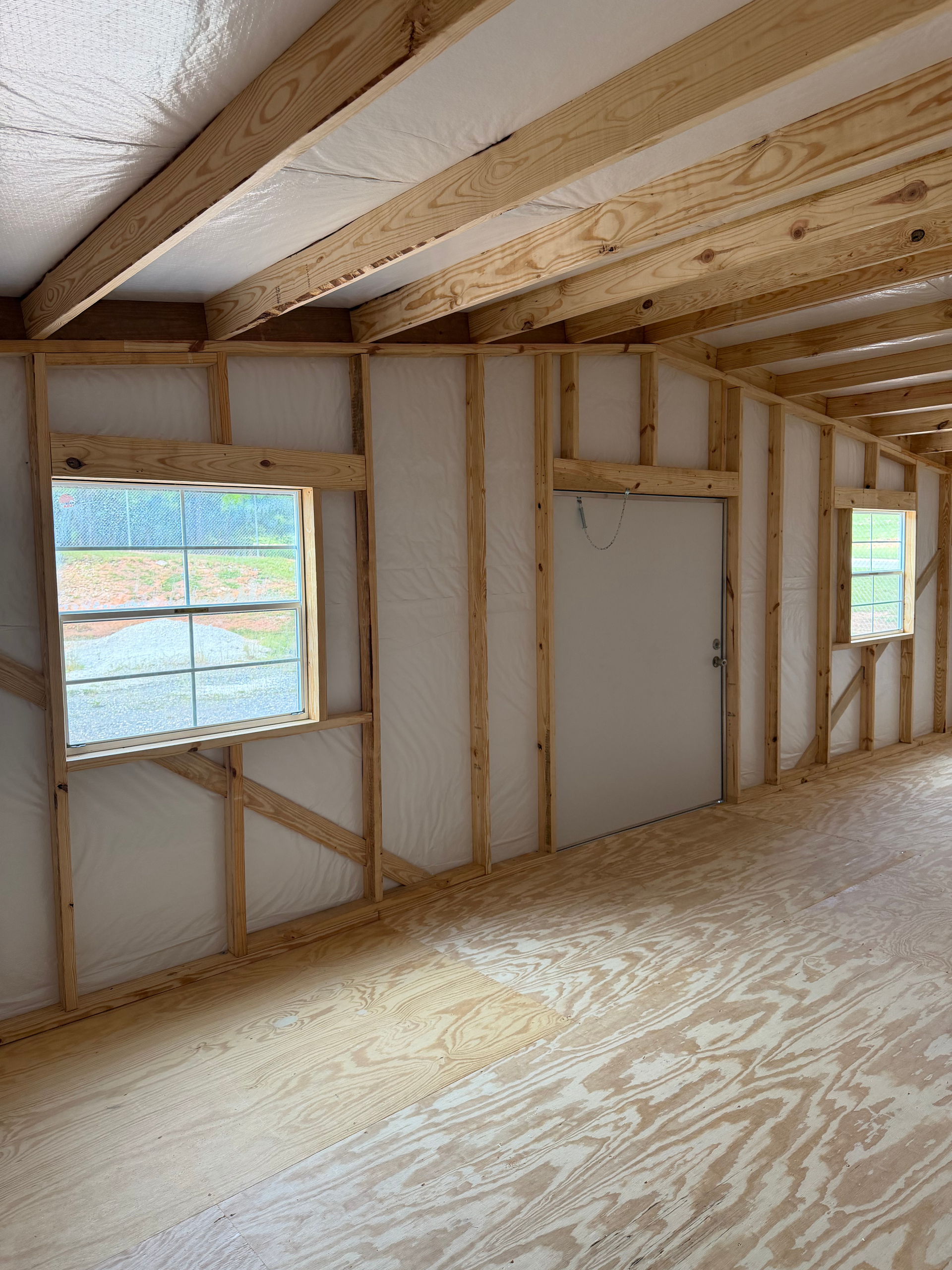 Unfinished room interior with exposed framing, insulation, plywood floor, and two windows.