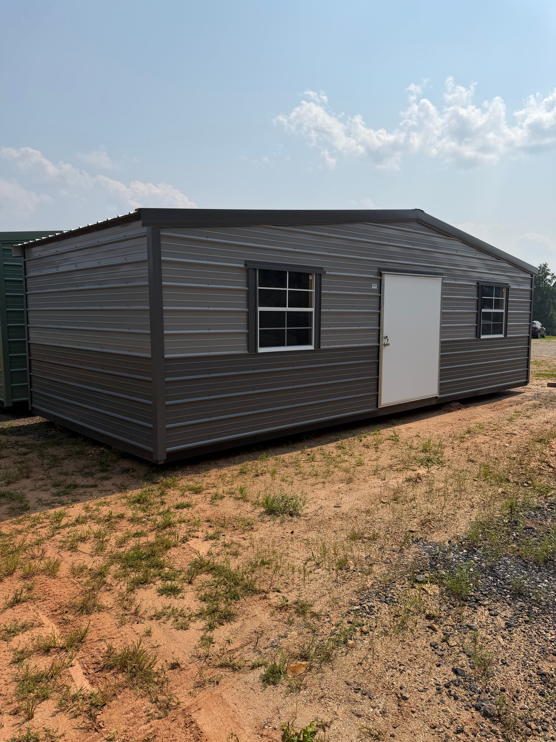 Gray metal shed with white door and small windows on a grassy dirt lot under a blue sky