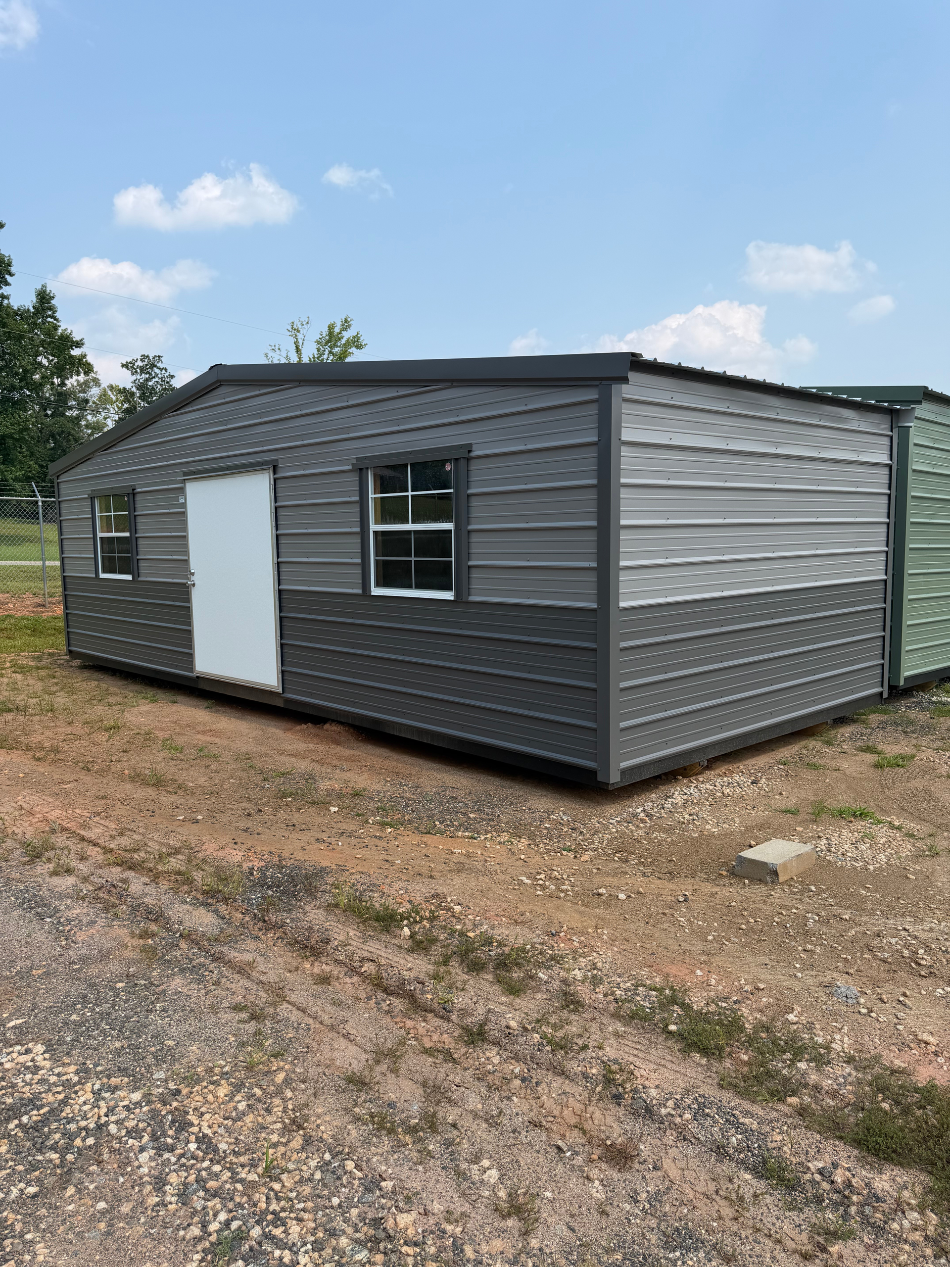 Gray metal shed with a white door and window, sitting on a gravel lot under a blue sky