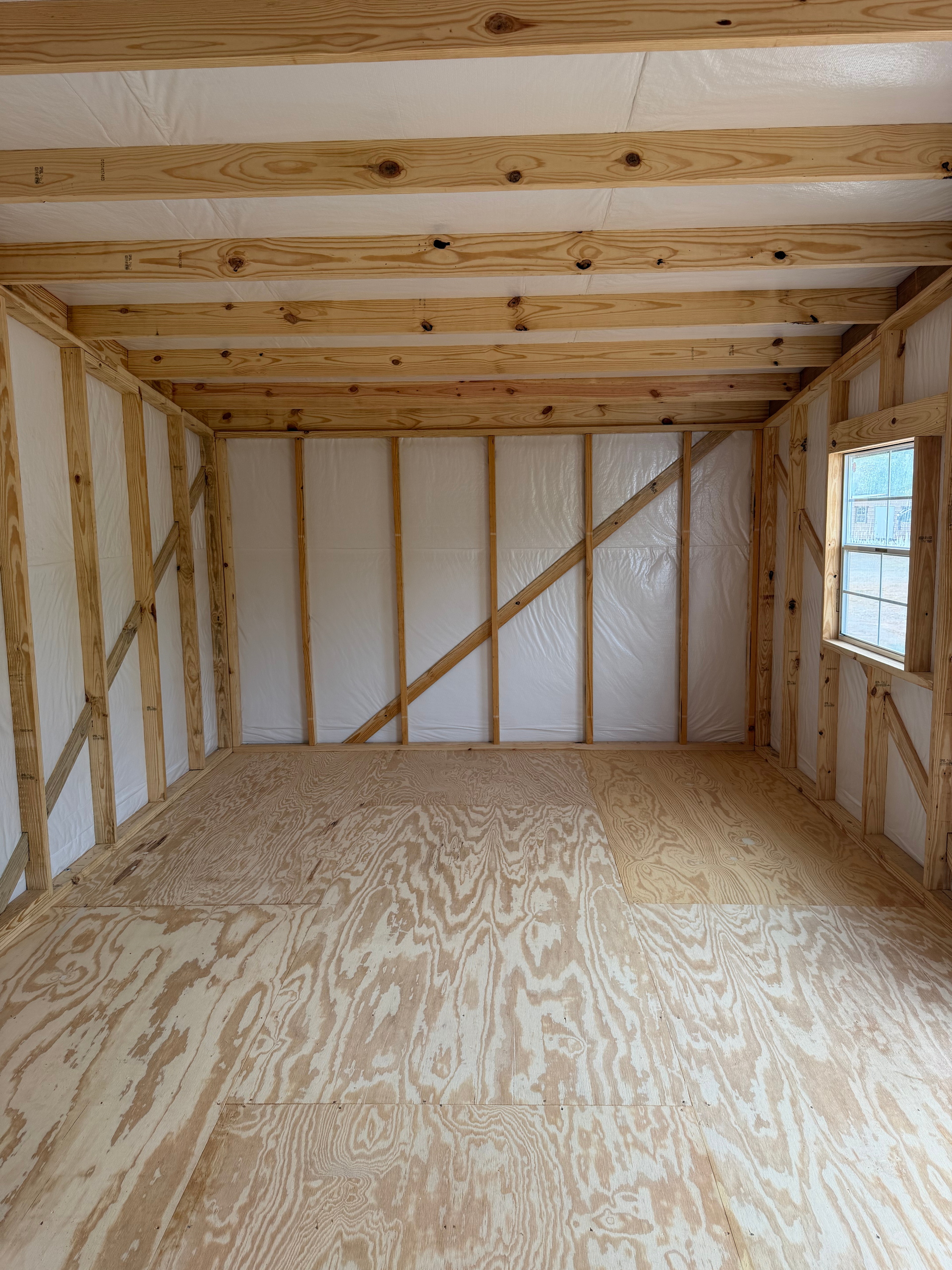Empty unfinished wooden room with plywood floor, exposed framing, and a small window on the right.