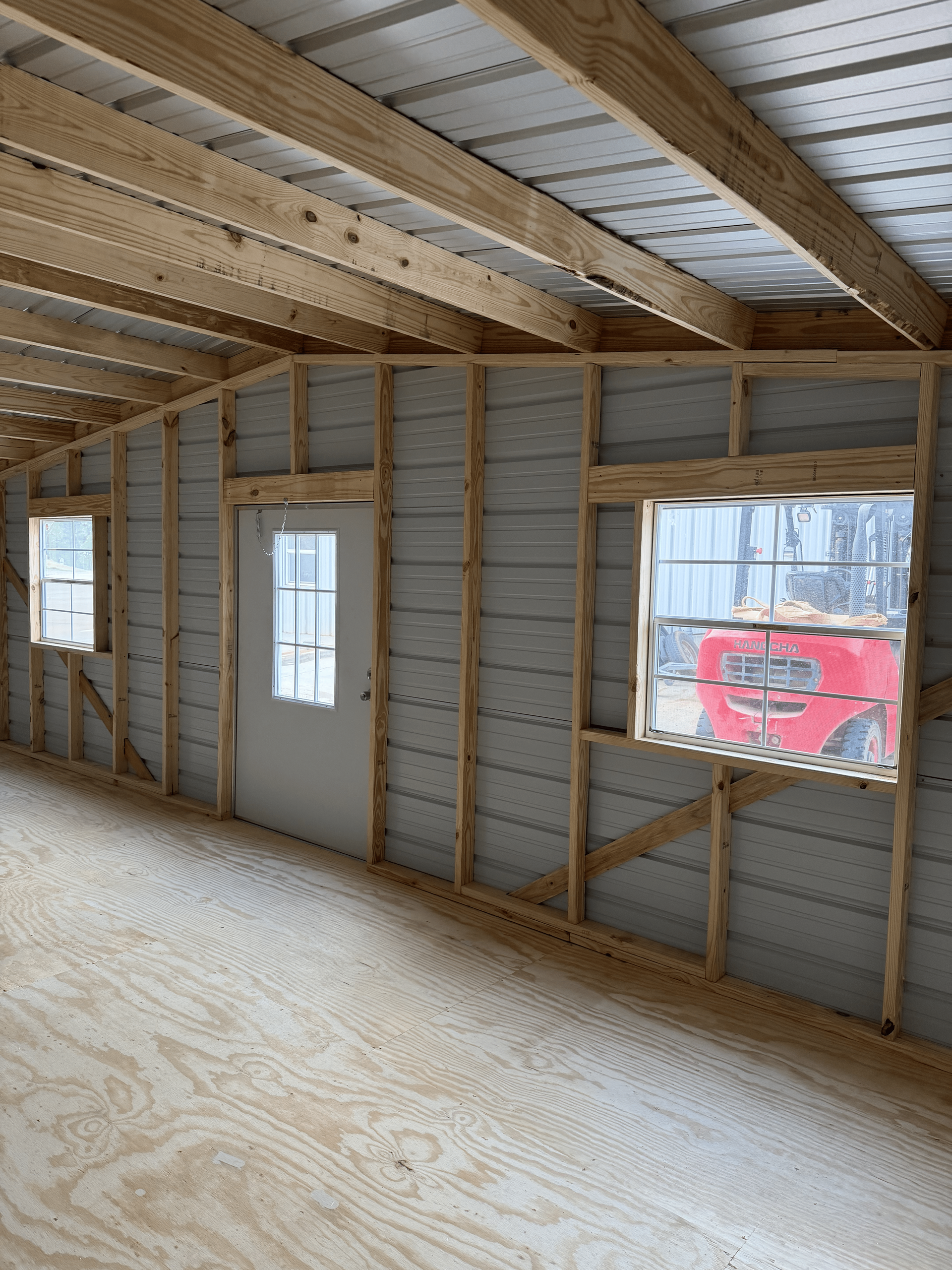Unfinished room with exposed studs, gray door, window openings, and plywood floor under construction