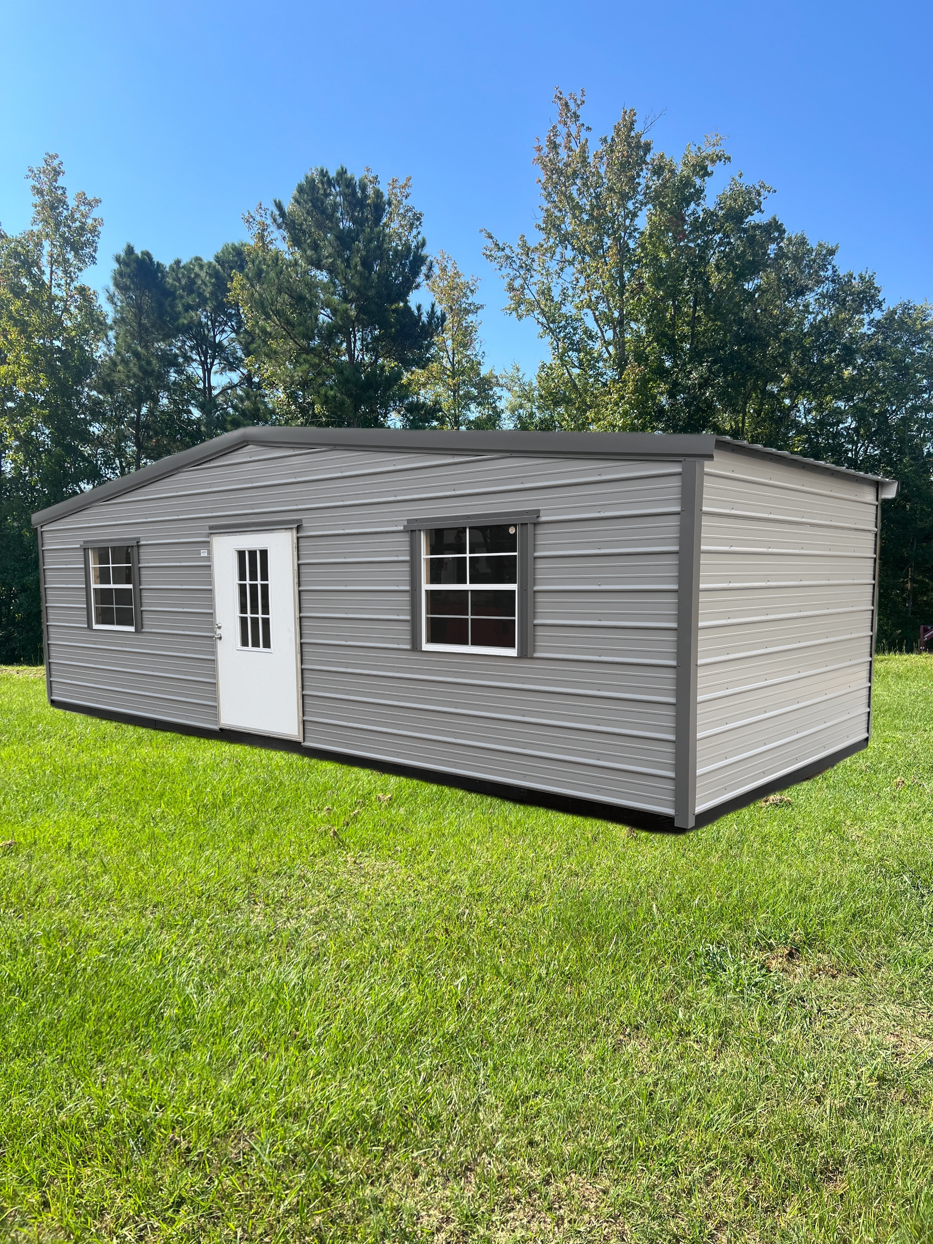 Small gray prefab cabin with white door and windows on a grassy lawn under a blue sky
