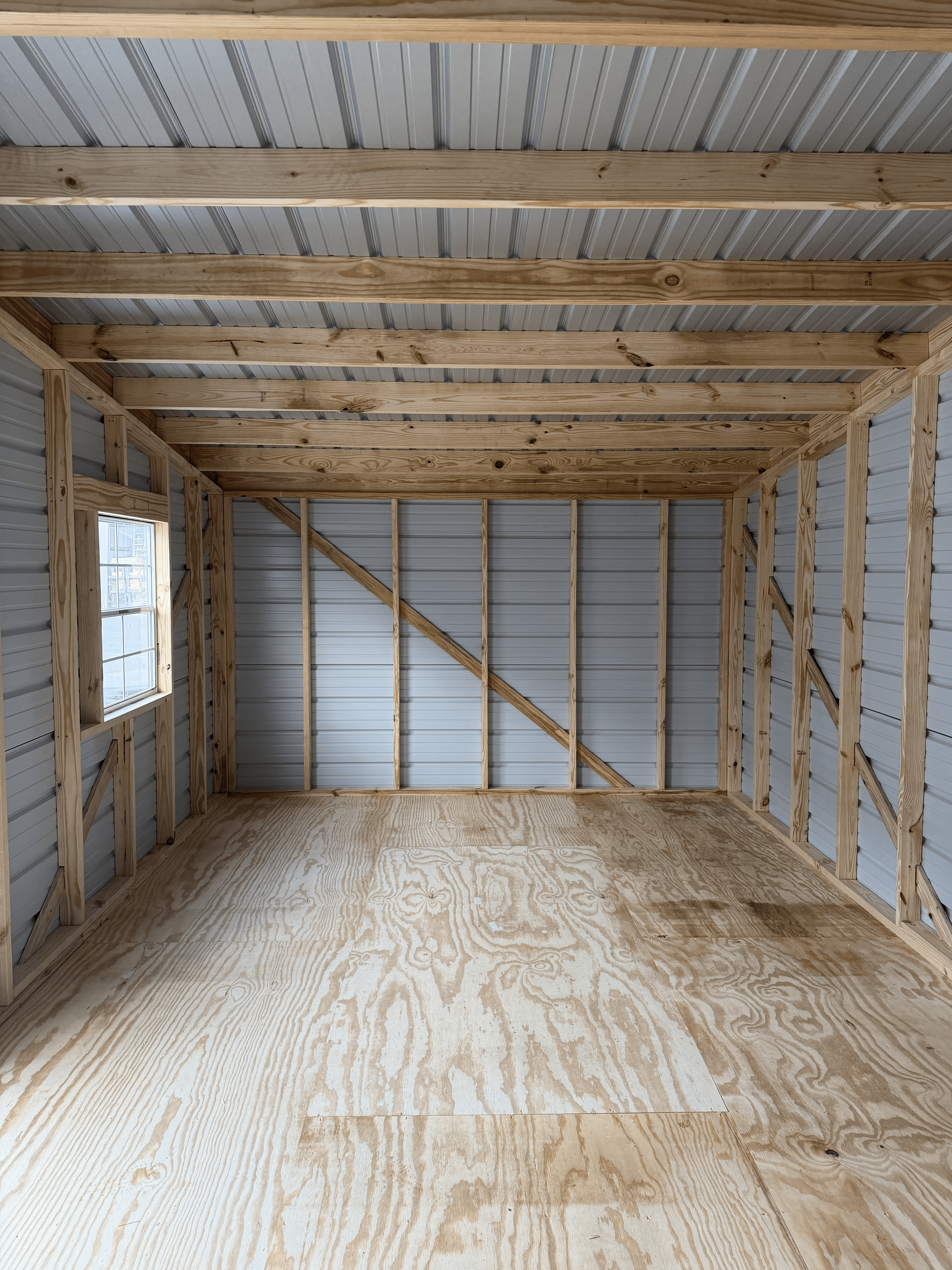 Unfinished shed interior with exposed wooden framing, plywood floor, and corrugated metal back wall.