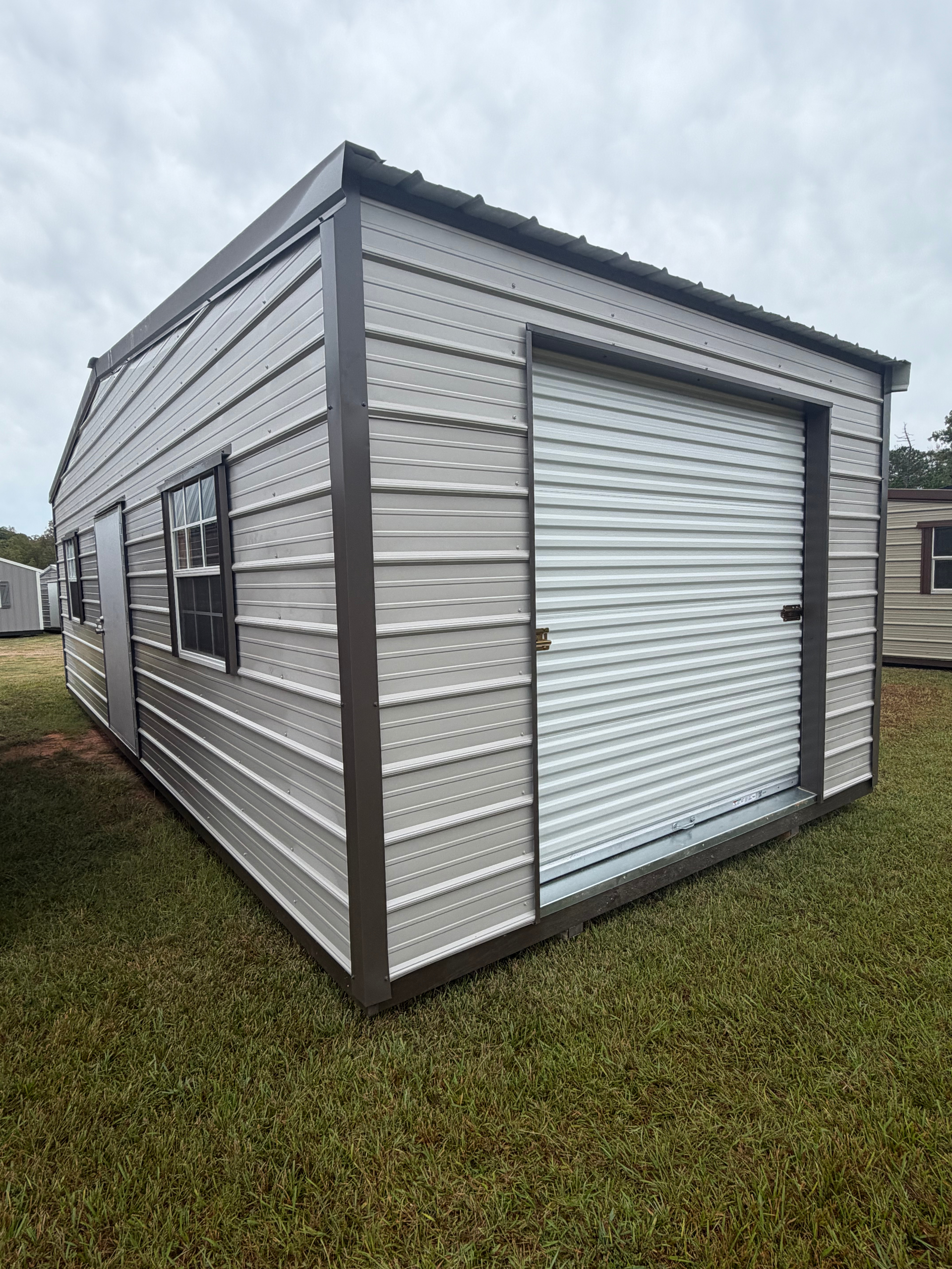 Small gray metal storage shed with a white roll-up door on a grassy lot
