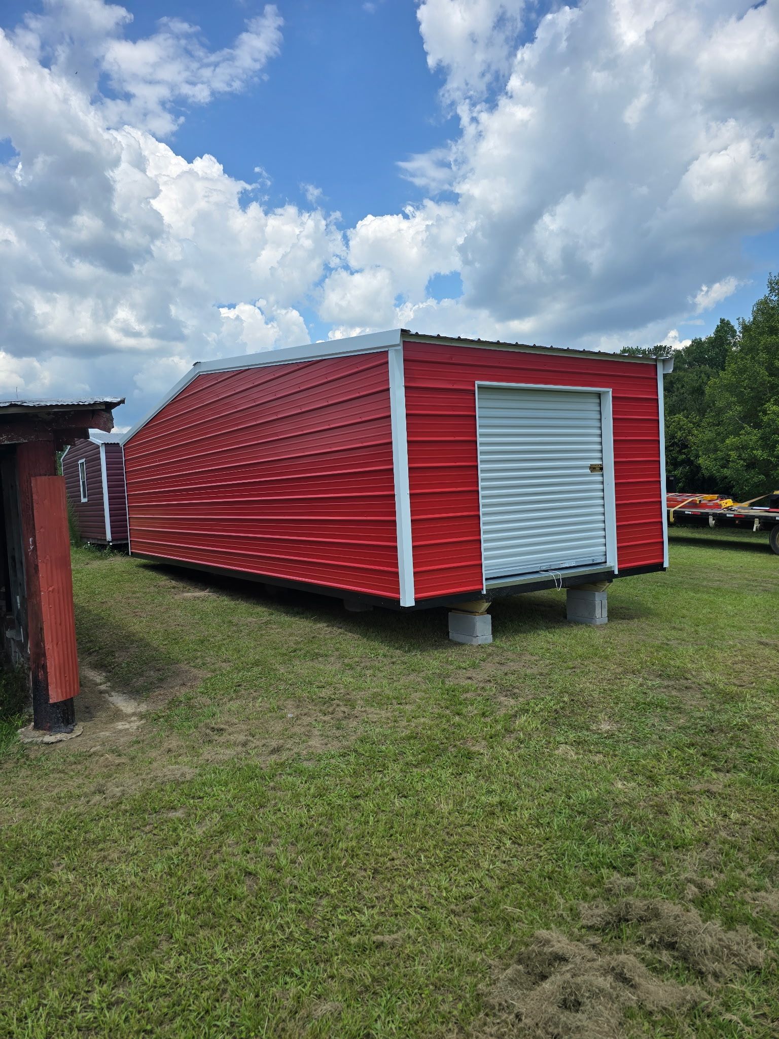 Red corrugated metal shed with a white roll-up door on grass under a partly cloudy sky