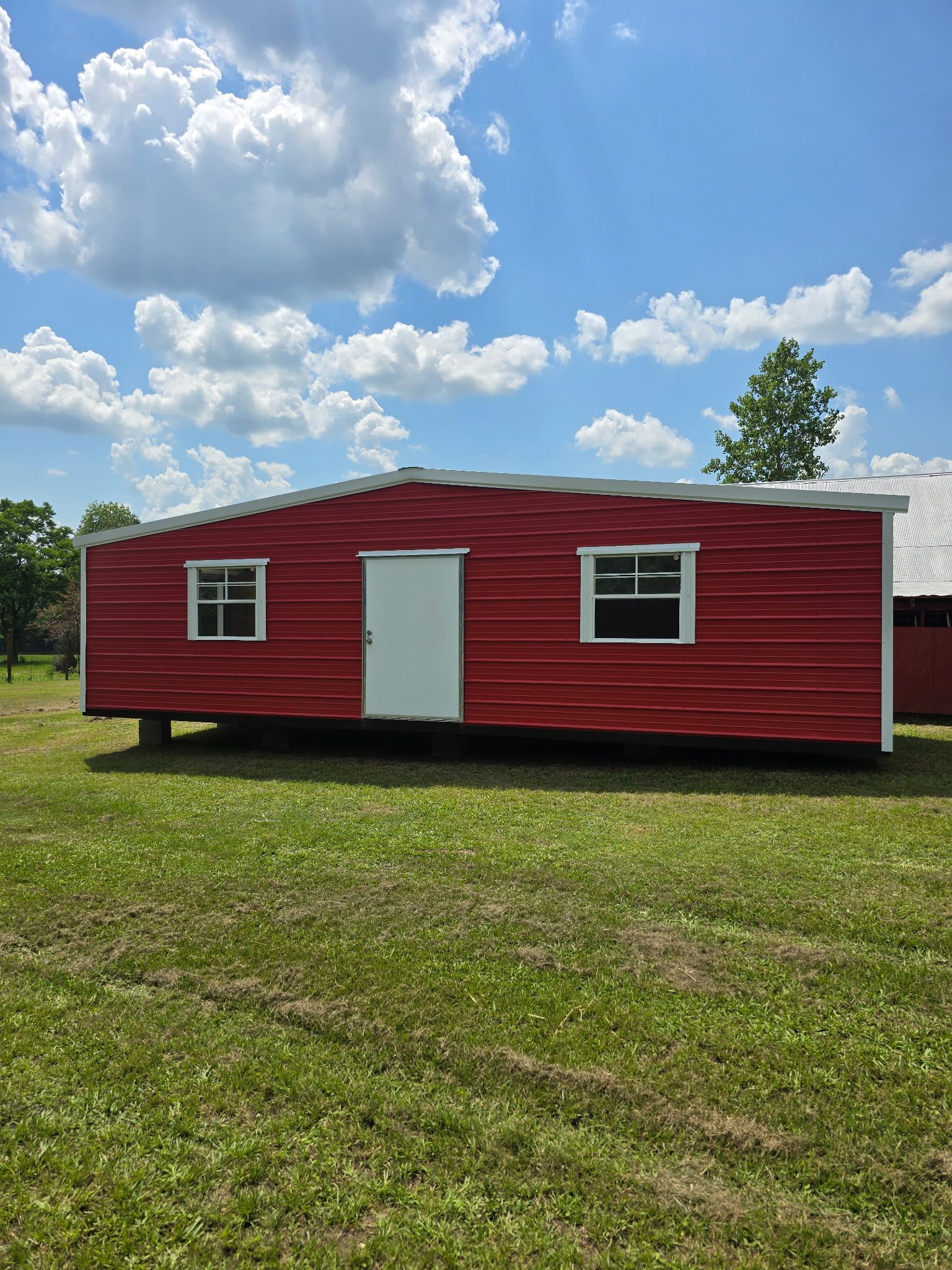 Red mobile home with white door and windows in a grassy field under a cloudy blue sky