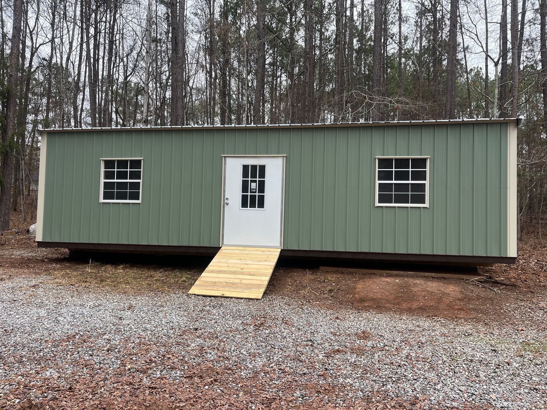 Green metal building with white door, two windows, and a ramp, set in a wooded area.